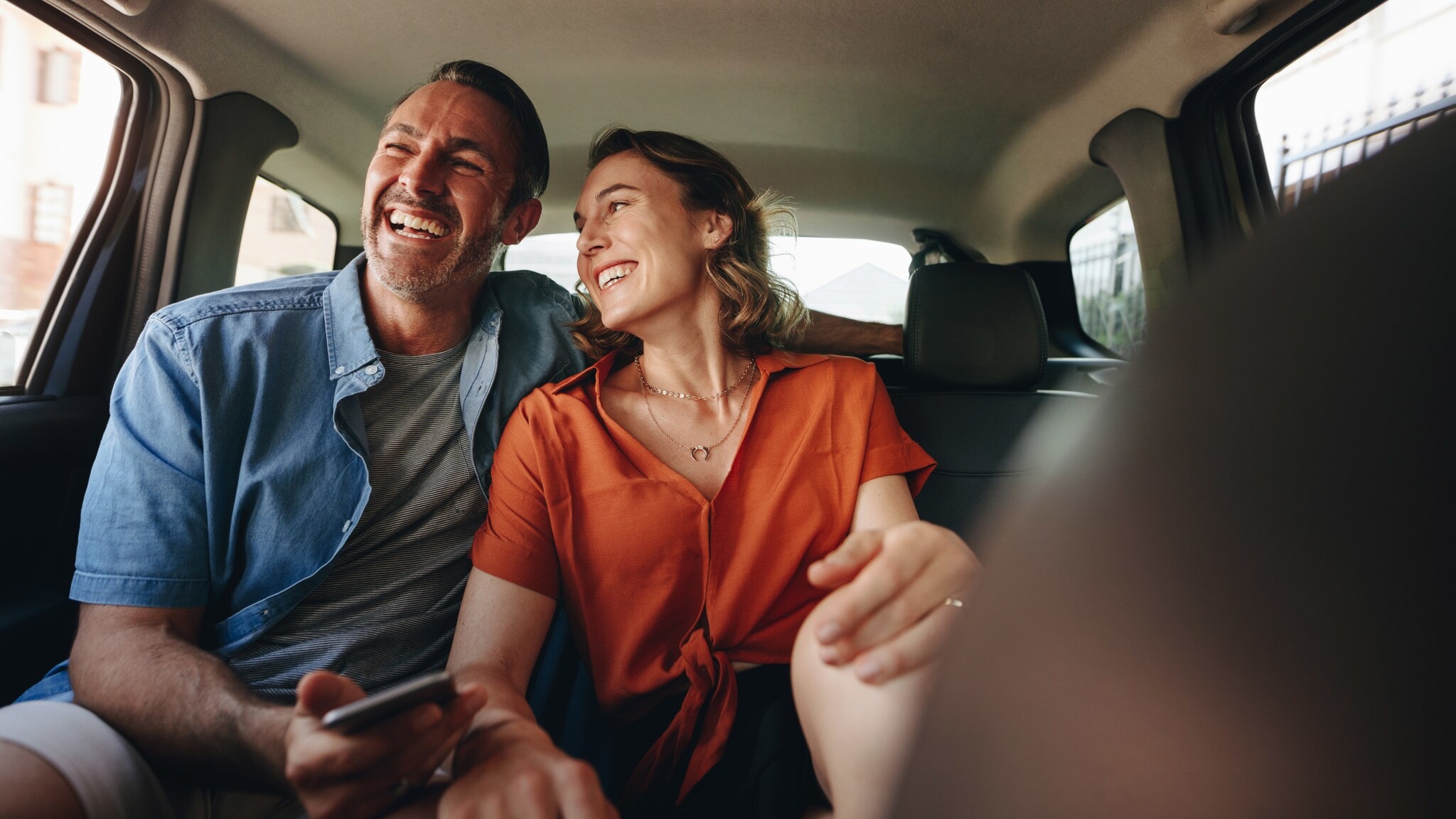 Ein fröhliches Paar sitzt auf der Rückbank eines Autos, er hält ein Smartphone in der Hand. Ein fröhliches Paar sitzt auf der Rückbank eines Autos, er hält ein Smartphone in der Hand.