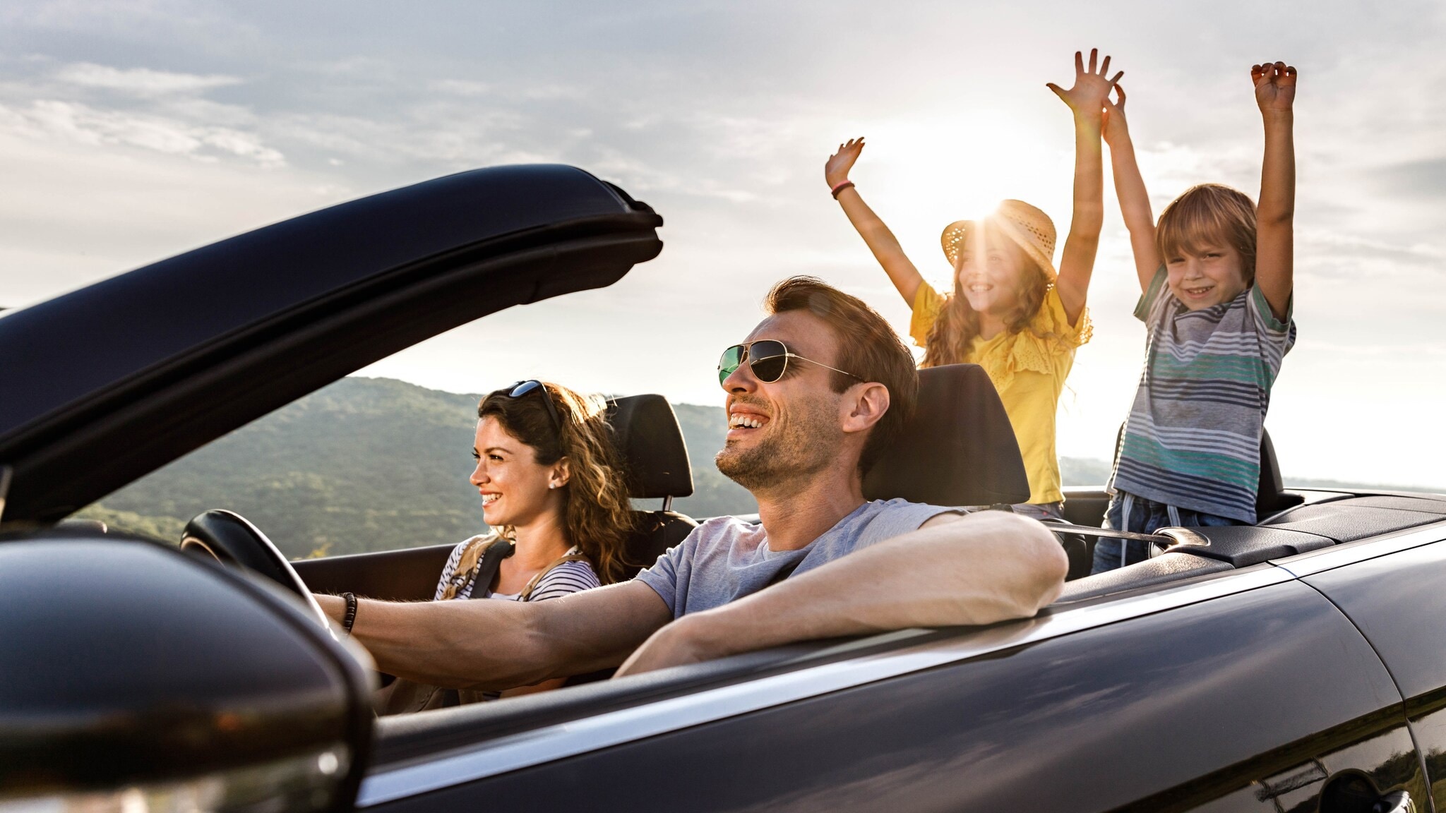 Eine Frau und ein Mann in einem offenen Cabriolet, auf den Rücksitzen zwei Kinder mit hochgestreckten Armen im Sonnenschein. Eine Frau und ein Mann in einem offenen Cabriolet, auf den Rücksitzen zwei Kinder mit hochgestreckten Armen im Sonnenschein.