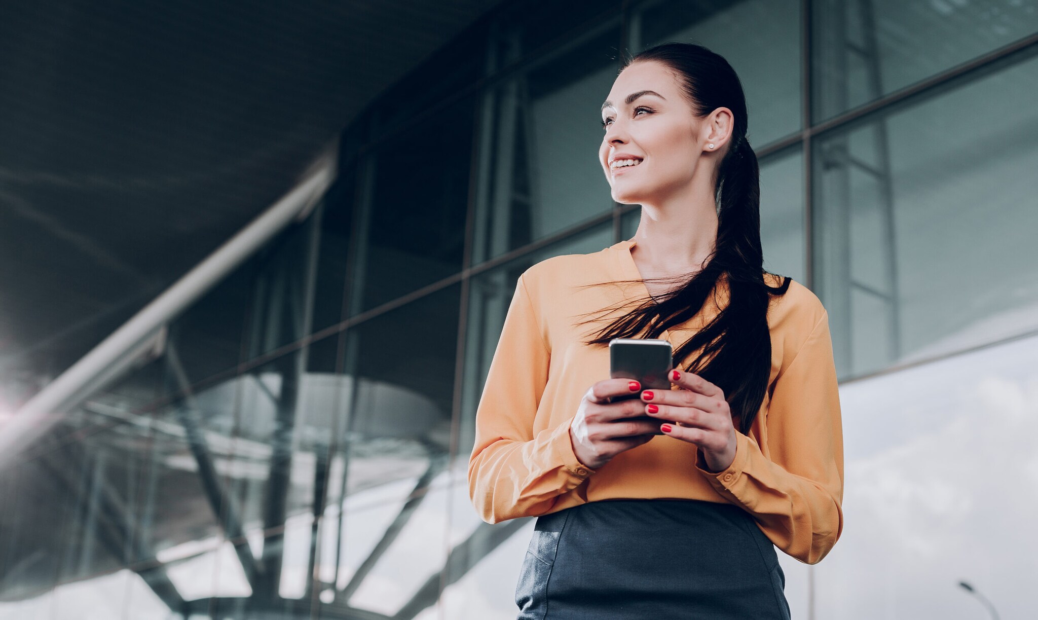 Eine junge, elegant gekleidete Frau mit einem Smartphone in den Händen steht lächelnd vor einem Flughafenterminal.