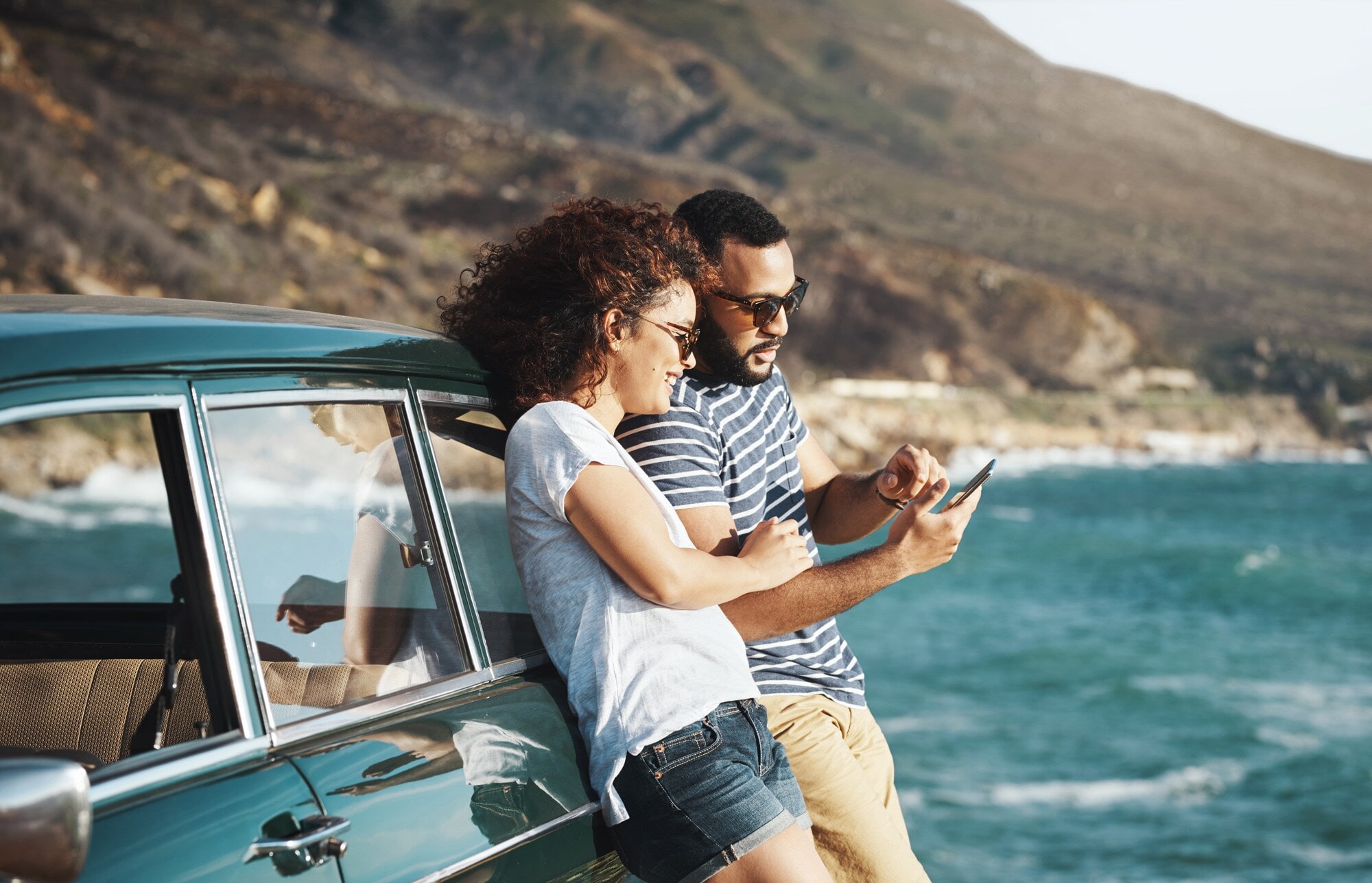 Ein Paar in Freizeitkleidung lehnt sich an ein Auto an einer Küste und schaut gemeinsam auf ein Smartphone Ein Paar in Freizeitkleidung lehnt sich an ein Auto an einer Küste und schaut gemeinsam auf ein Smartphone