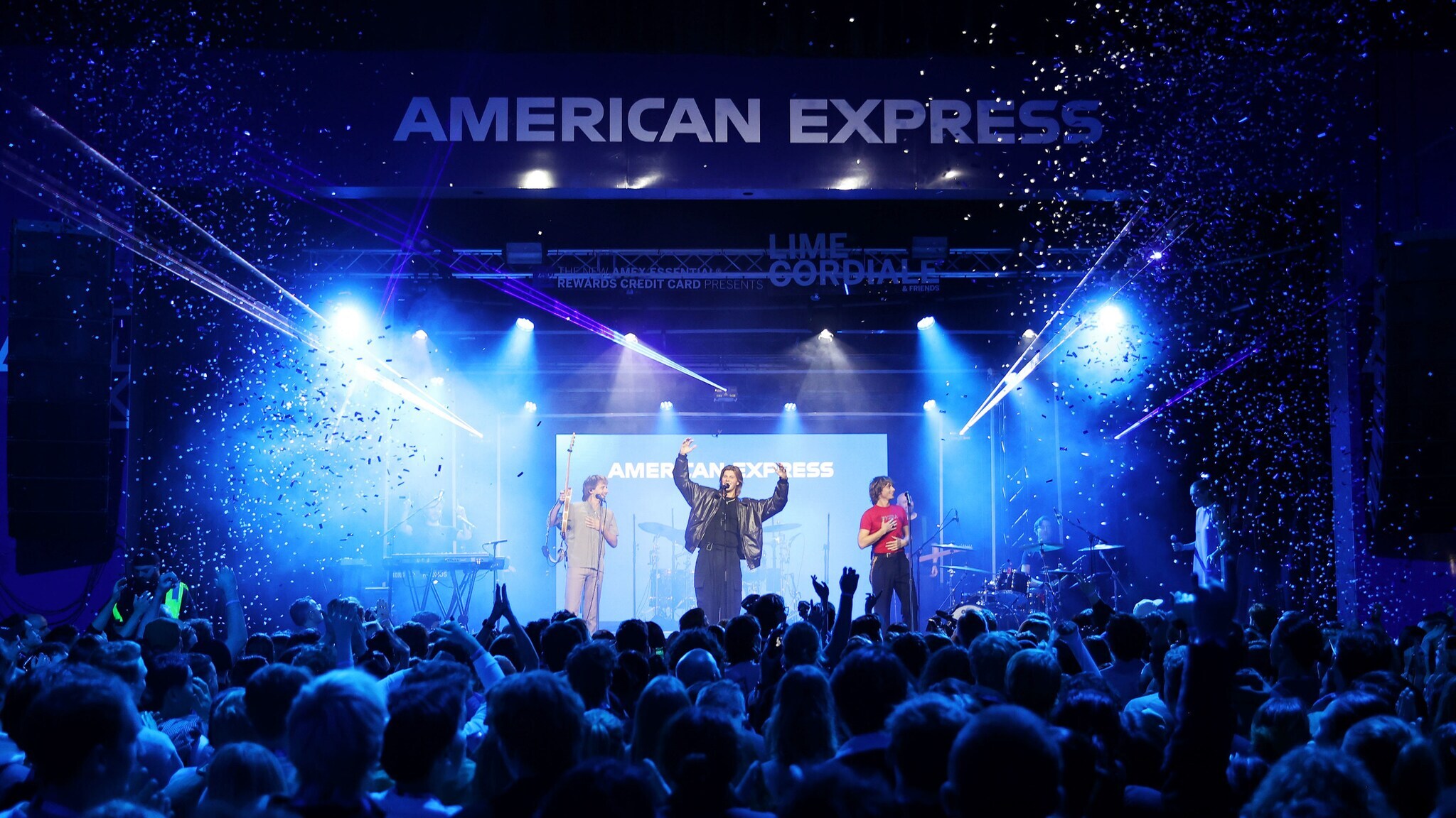 Eine Band auf einer blau beleuchteten Bühne mit American Express Schriftzug vor Publikum.