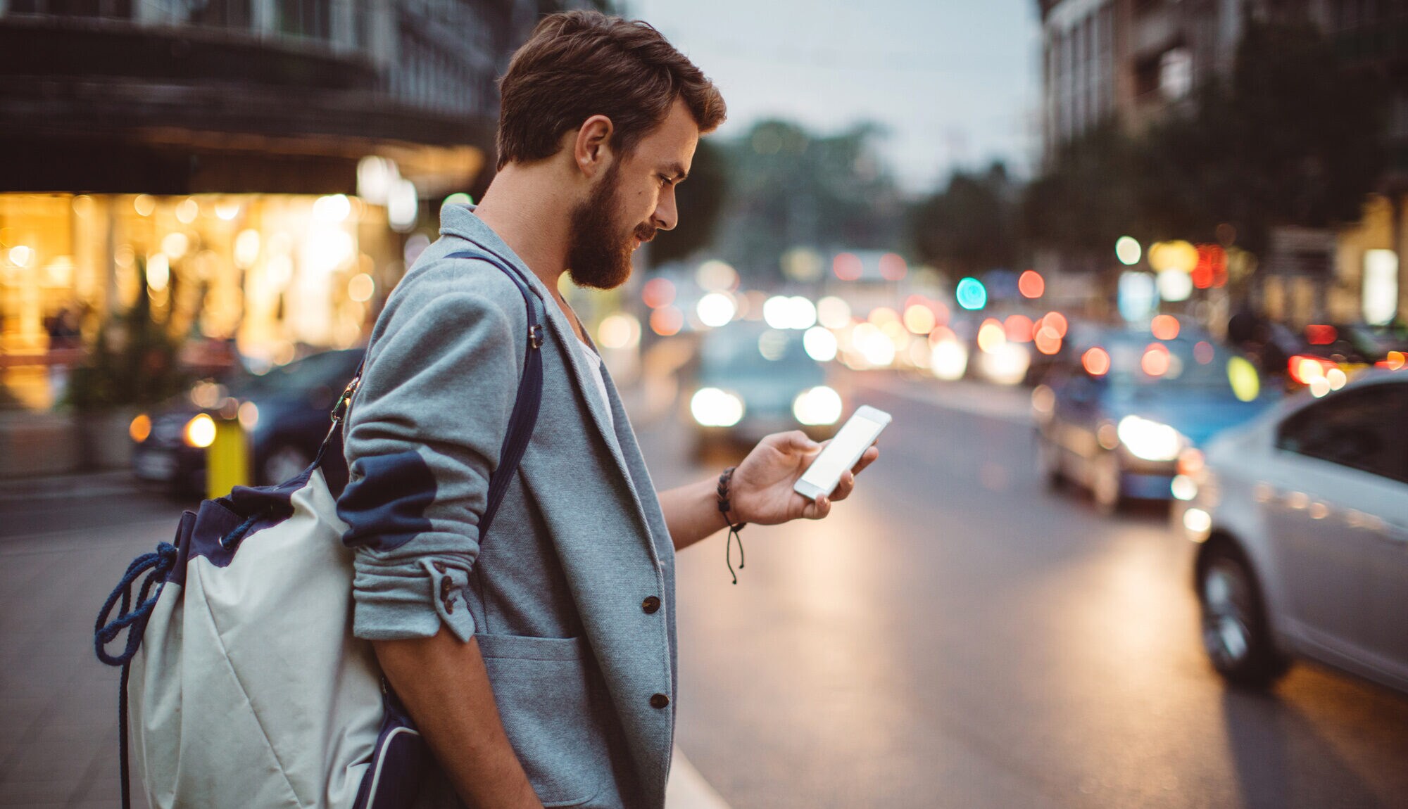 Ein junger Mann mit Rucksack steht an einer befahrenen Straße und schaut auf sein Smartphone