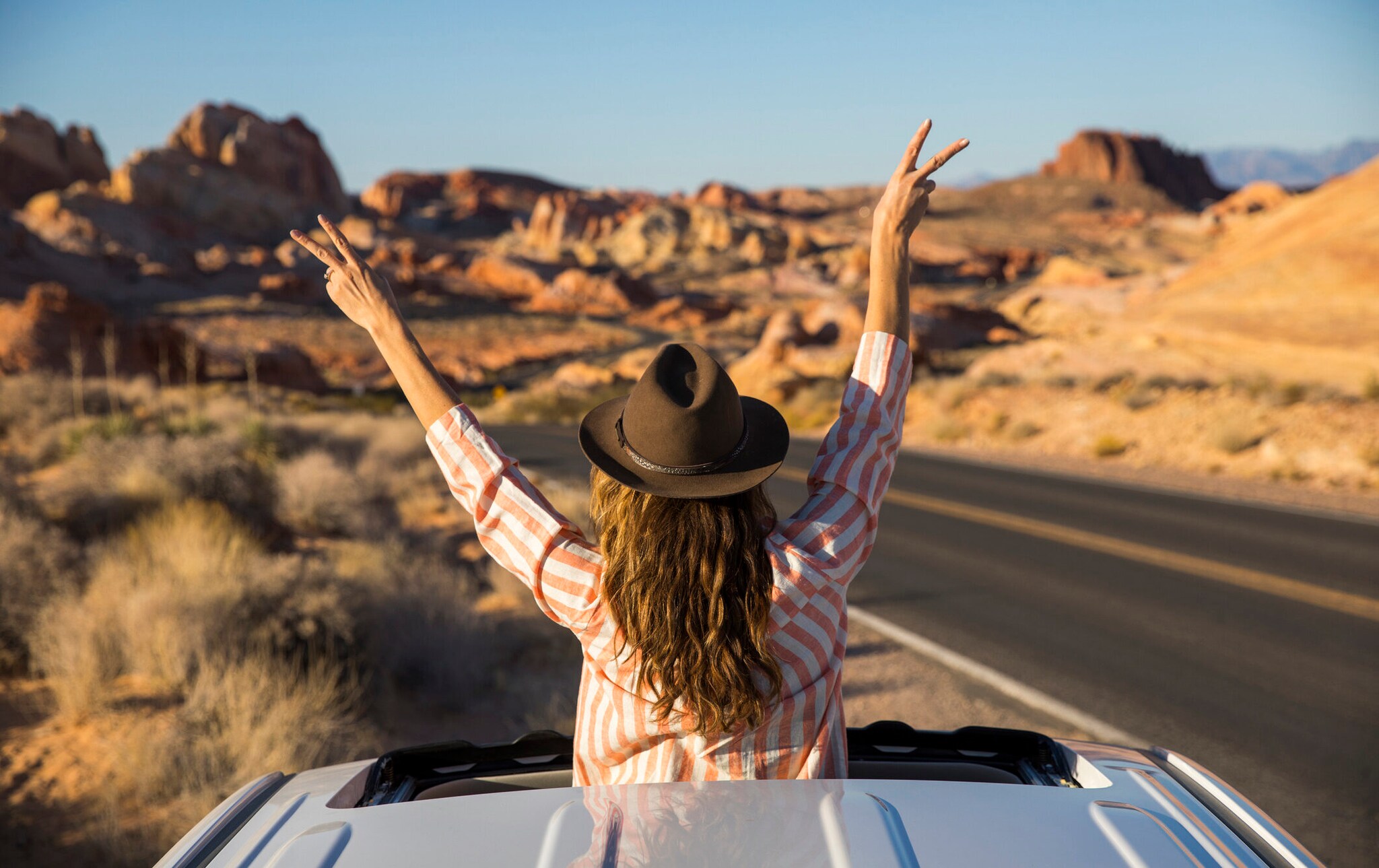 Rückansicht einer Frau mit Hut, die auf einer Reise durch eine Wüstenlandschaft mit dem Oberkörper aus dem Schiebedach eines Mietwagens schaut und die Hände in die Luft streckt