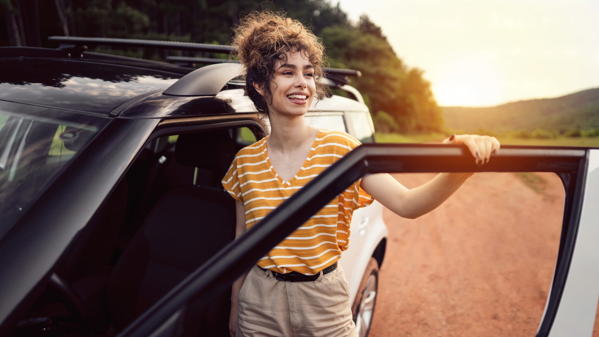 Eine lächelnde Frau steht in der geöffneten Tür ihres Autos auf einer Landstraße. Eine lächelnde Frau steht in der geöffneten Tür ihres Autos auf einer Landstraße.