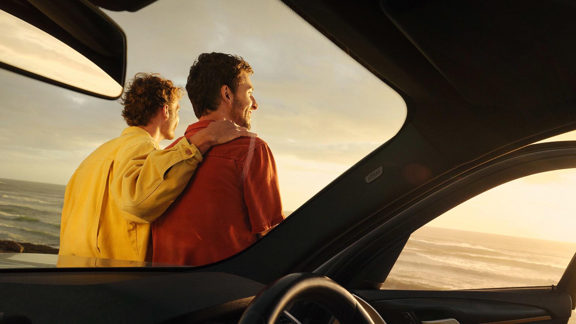 Blick aus dem Inneren eines Autos auf die Rückansicht eines Paares, das an einem Auto lehnt, vor Meerespanorama.