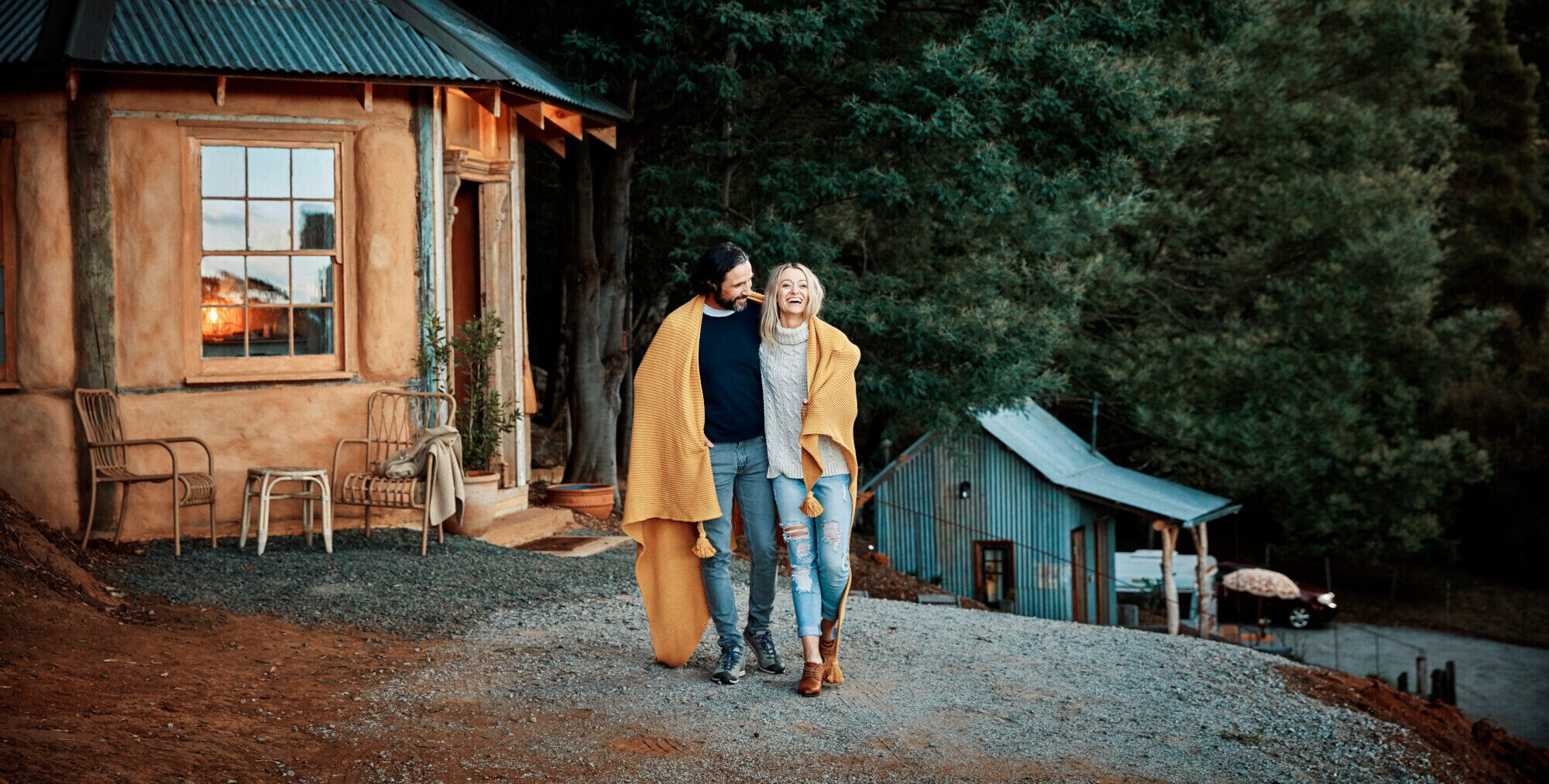 Ein glückliches, stylishes Paar steht in eine Decke gekuschelt vor einer gemütlichen Holzhütte in einem Waldgebiet