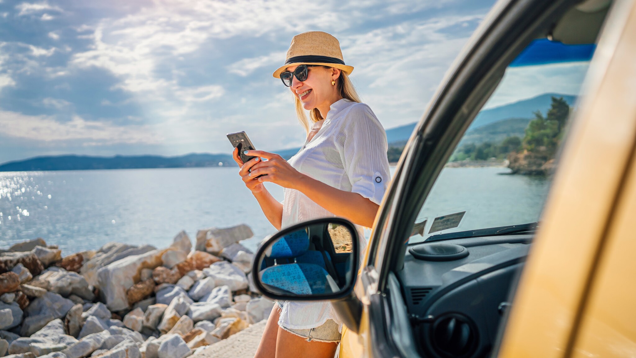 Eine Frau mit Sonnenbrille und Strohhut lehnt lächelnd an einem Auto und schaut auf ihr Smartphone an einer Küste bei Sonnenschein.