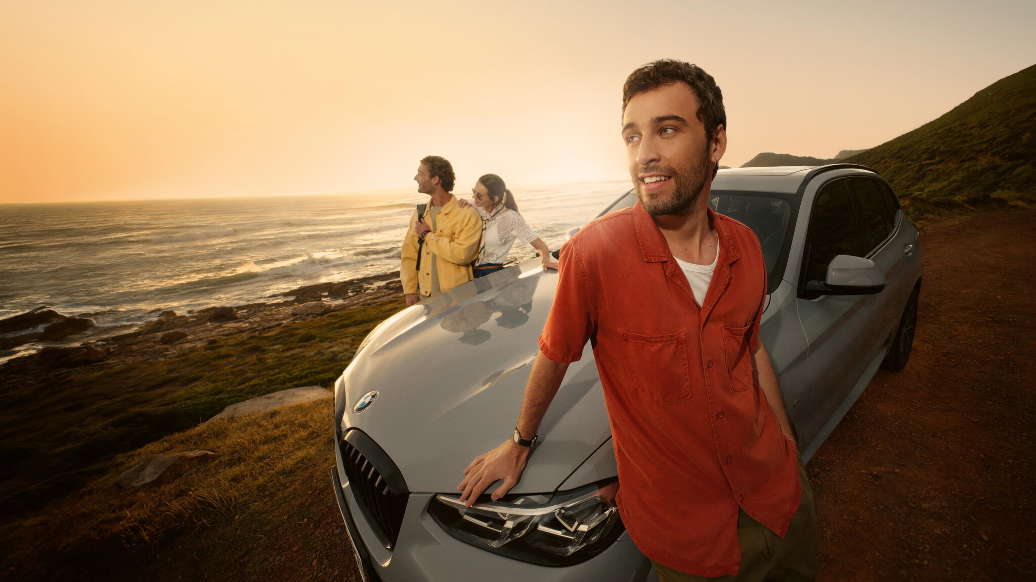 Ein junger Mann lehnt an der Motorhaube eines Autos an einer Küste, hinter dem Auto stehen zwei weitere Personen vor Meerespanorama.