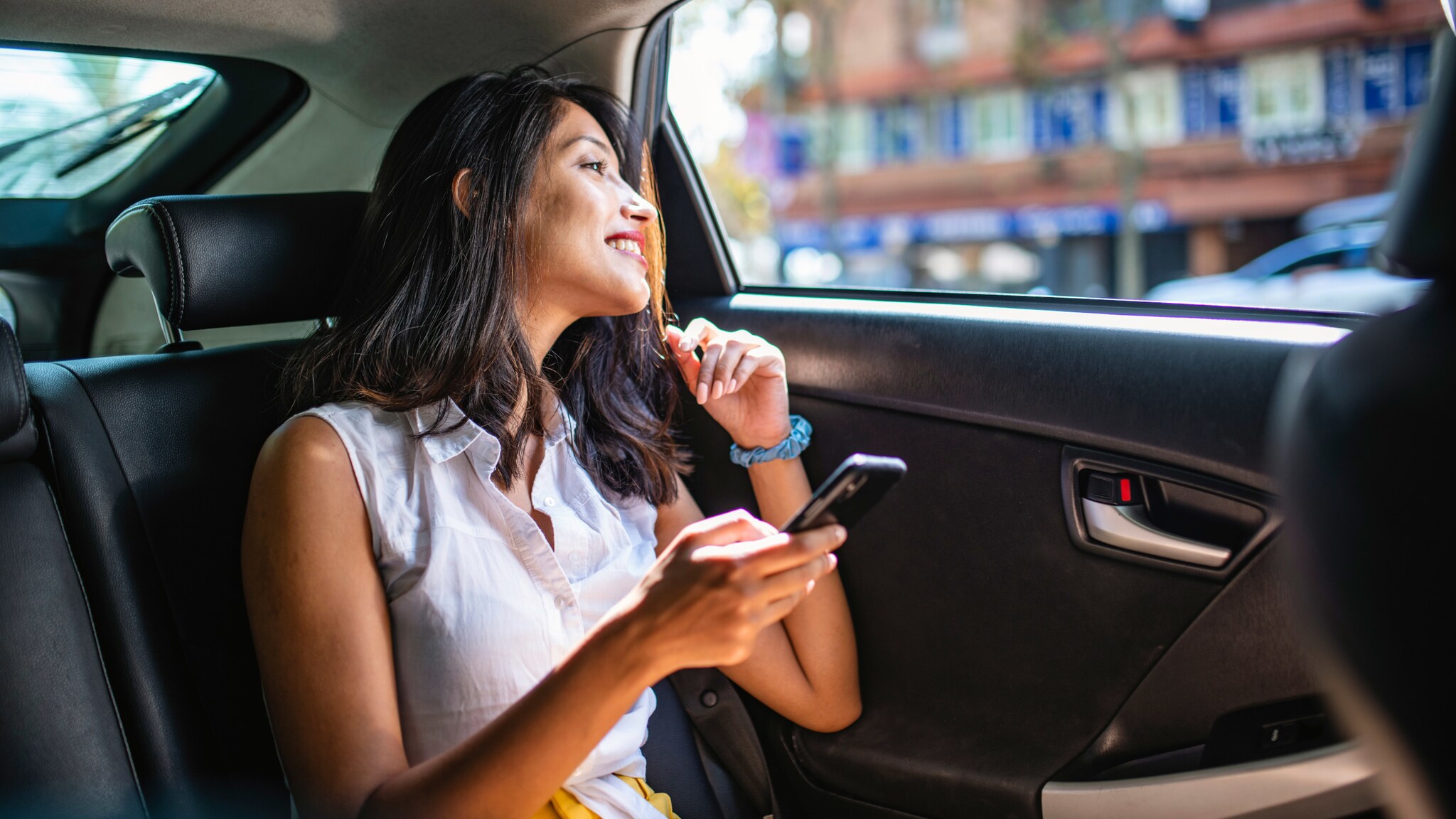 Eine gut gelaunte Frau sitzt auf der Rückbank eines Autos und schaut aus dem geöffneten Fenster, in ihrer Hand hält sie ein Smartphone.