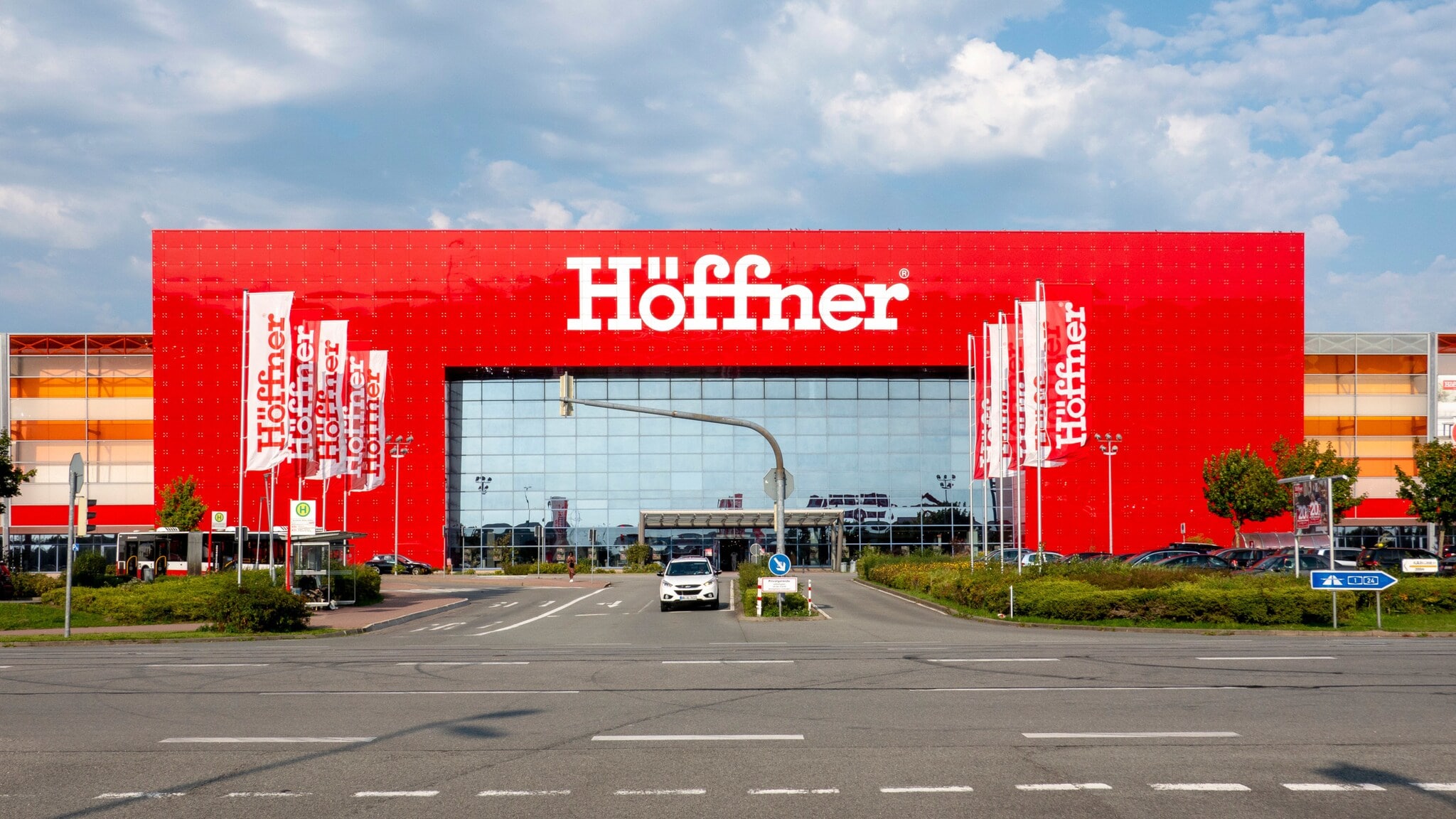 Großes, freistehendes Gebäude mit Schriftzug „Höffner“ auf roter Fassade hinter einer Straßenkreuzung mit rot-weißen Fahnen. Großes, freistehendes Gebäude mit Schriftzug „Höffner“ auf roter Fassade hinter einer Straßenkreuzung mit rot-weißen Fahnen.