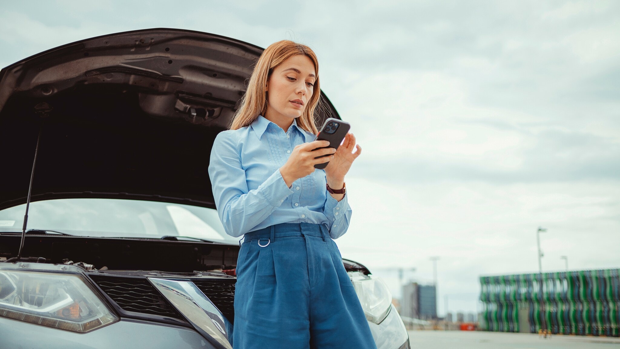 Eine Frau lehnt an einem Auto mit geöffneter Kühlerhaube und bedient ihr Smartphone. Eine Frau lehnt an einem Auto mit geöffneter Kühlerhaube und bedient ihr Smartphone.