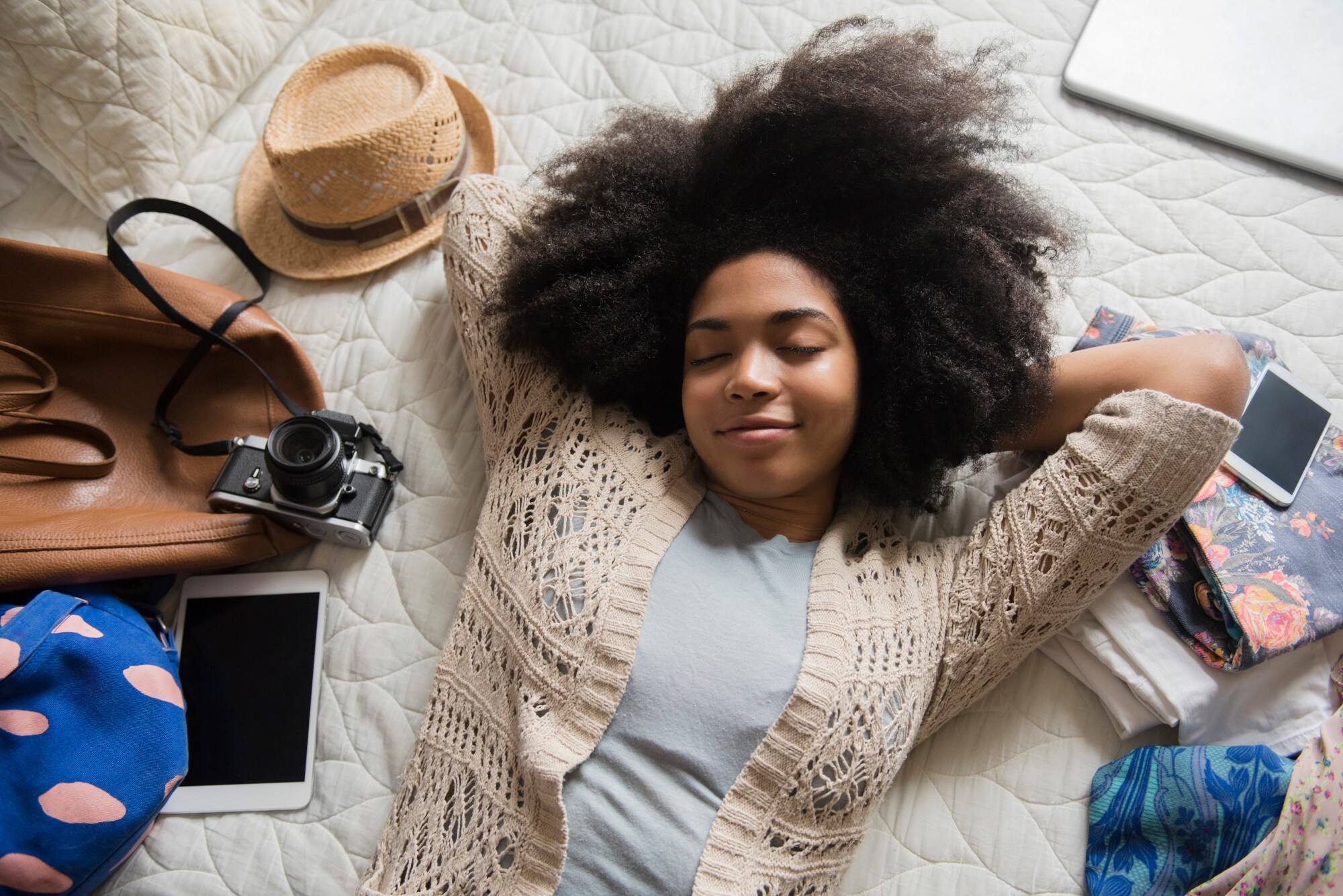 Aufsicht einer jungen Frau, die mit geschlossenen Augen zwischen Reiseutensilien auf einem Bett entspannt Aufsicht einer jungen Frau, die mit geschlossenen Augen zwischen Reiseutensilien auf einem Bett entspannt