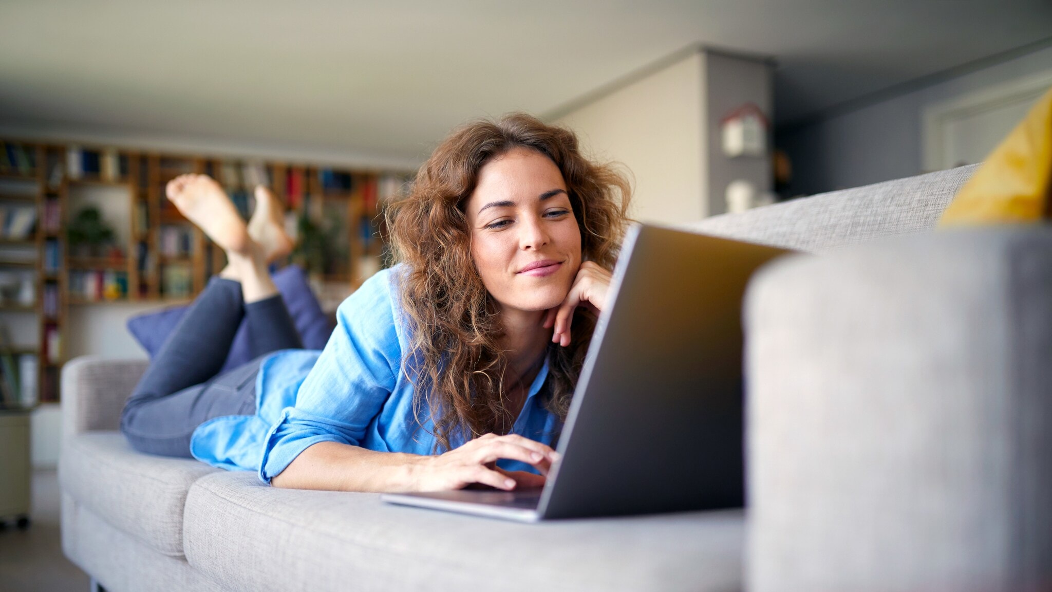 Eine Frau liegt bäuchlings auf einem Sofa in einem Wohnzimmer und schaut lächelnd auf einen Laptop vor ihr. Eine Frau liegt bäuchlings auf einem Sofa in einem Wohnzimmer und schaut lächelnd auf einen Laptop vor ihr.