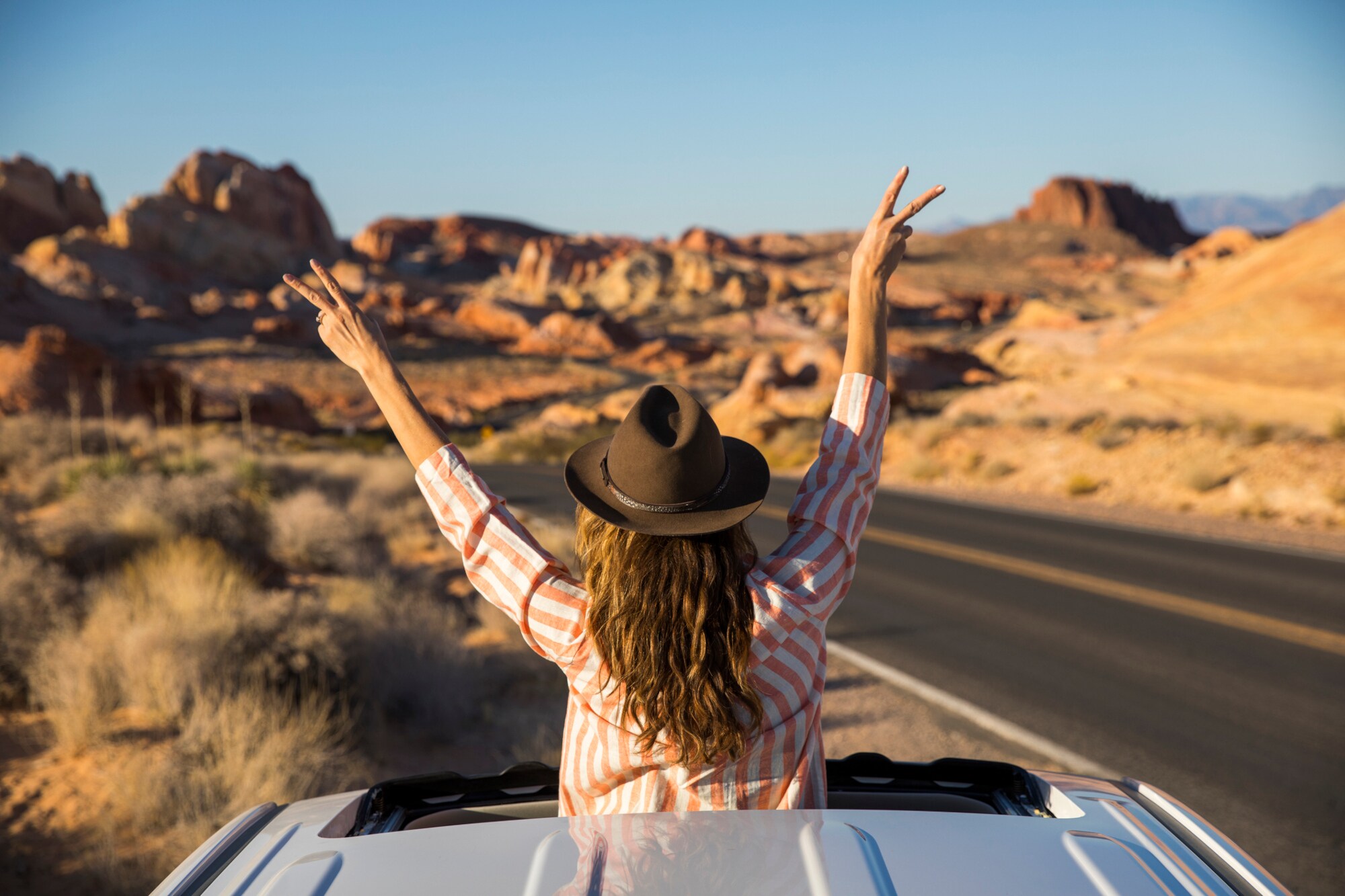 Eine junge Frau ragt mit dem Oberkörper aus dem Schiebedach eines Autos auf einer Straße in einer Wüstenlandschaft und winkt spontan mit einem Hut in die Luft
