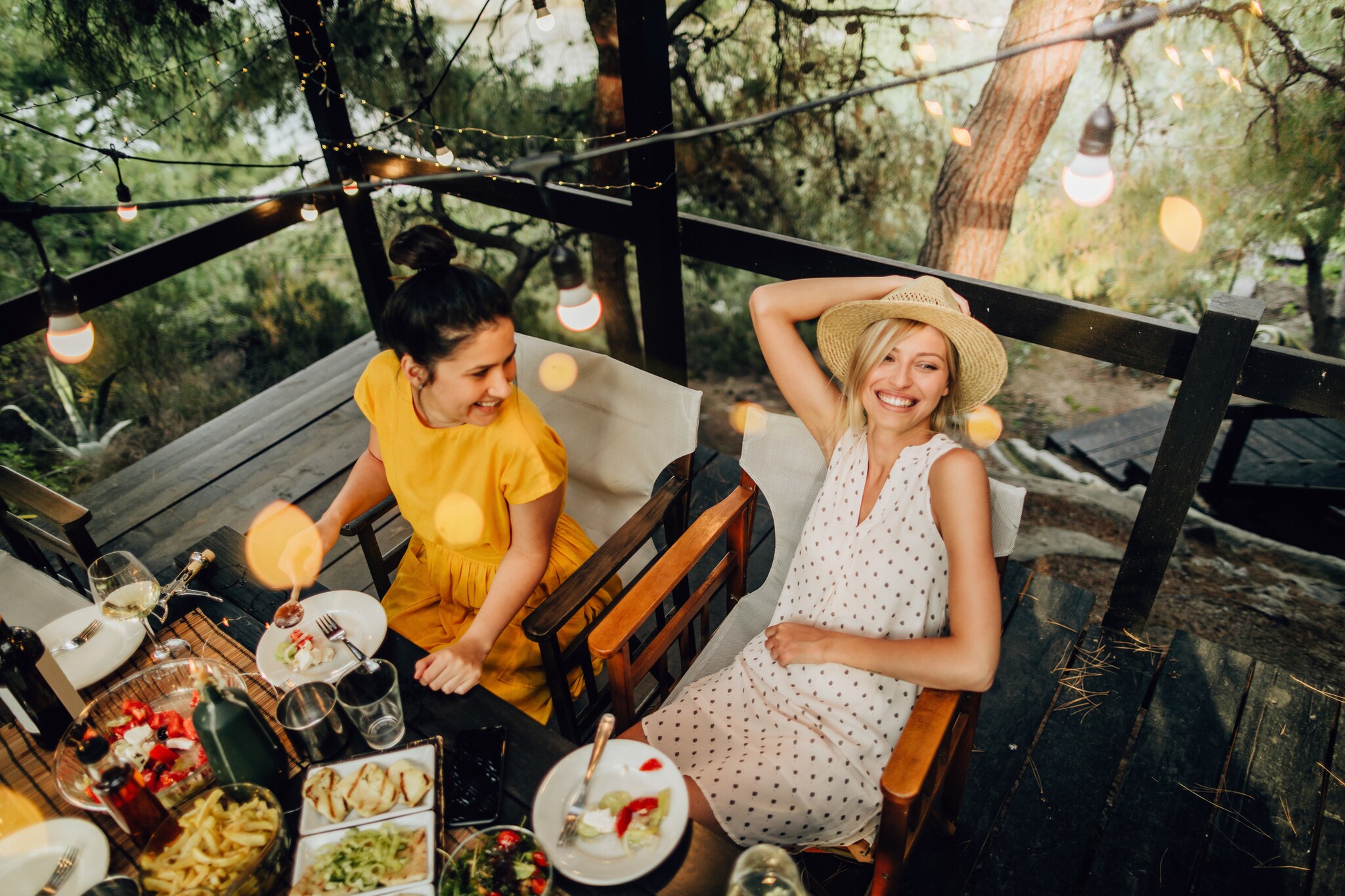 Zwei Frauen sitzen gut gelaunt beim sommerlichen Outdoor-Dinner an einem Tisch auf einer Holzterrasse Zwei Frauen sitzen gut gelaunt beim sommerlichen Outdoor-Dinner an einem Tisch auf einer Holzterrasse