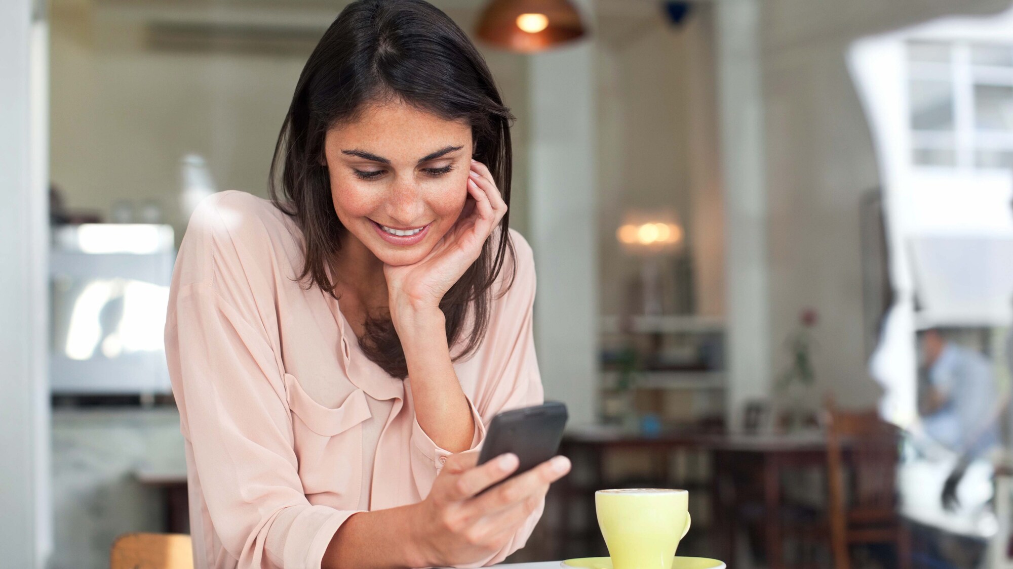 Eine junge Frau an einem Tisch in einem Café schaut lächelnd auf ihr Smartphone