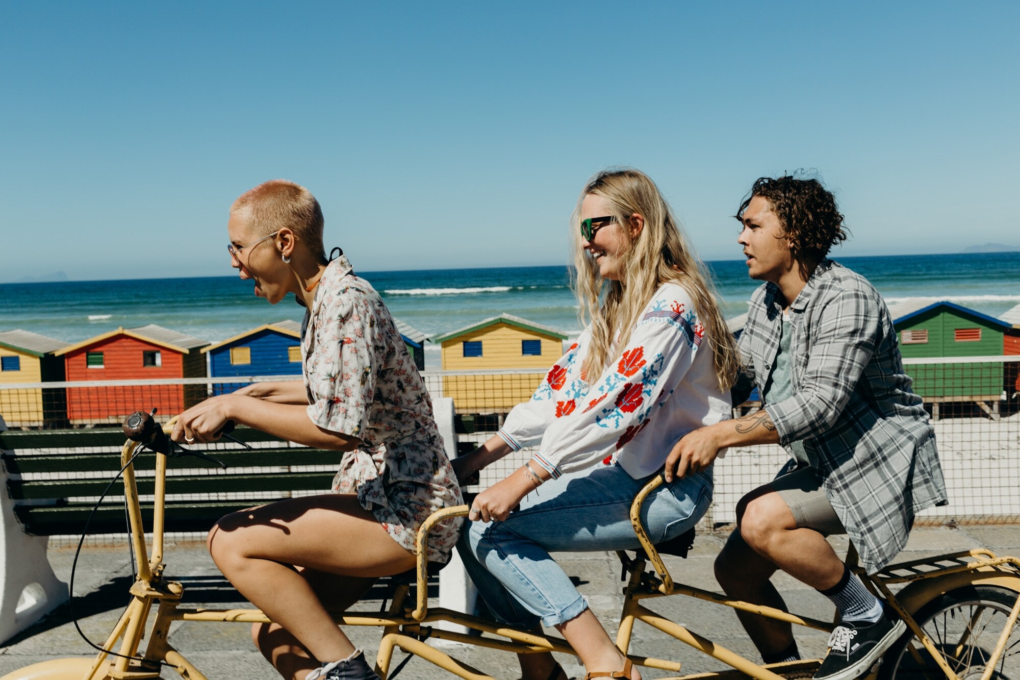 Drei junge Student:innen fahren auf einem Tridem im Sonnenschein am Meer entlang Drei junge Student:innen fahren auf einem Tridem im Sonnenschein am Meer entlang