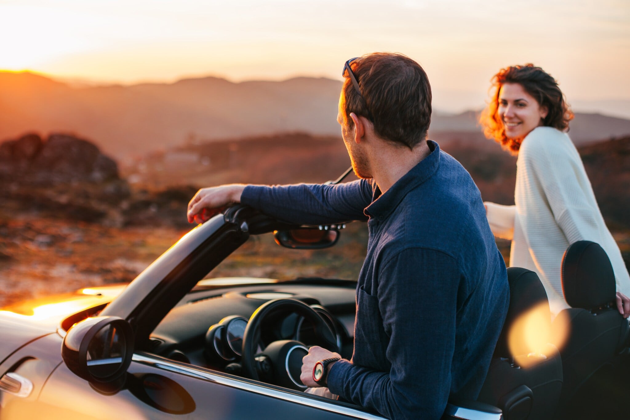 Rückansicht eines Paares auf Reise, das aus einem Cabriolet in eine Wüstenlandschaft bei Sonnenuntergang schaut Rückansicht eines Paares auf Reise, das aus einem Cabriolet in eine Wüstenlandschaft bei Sonnenuntergang schaut