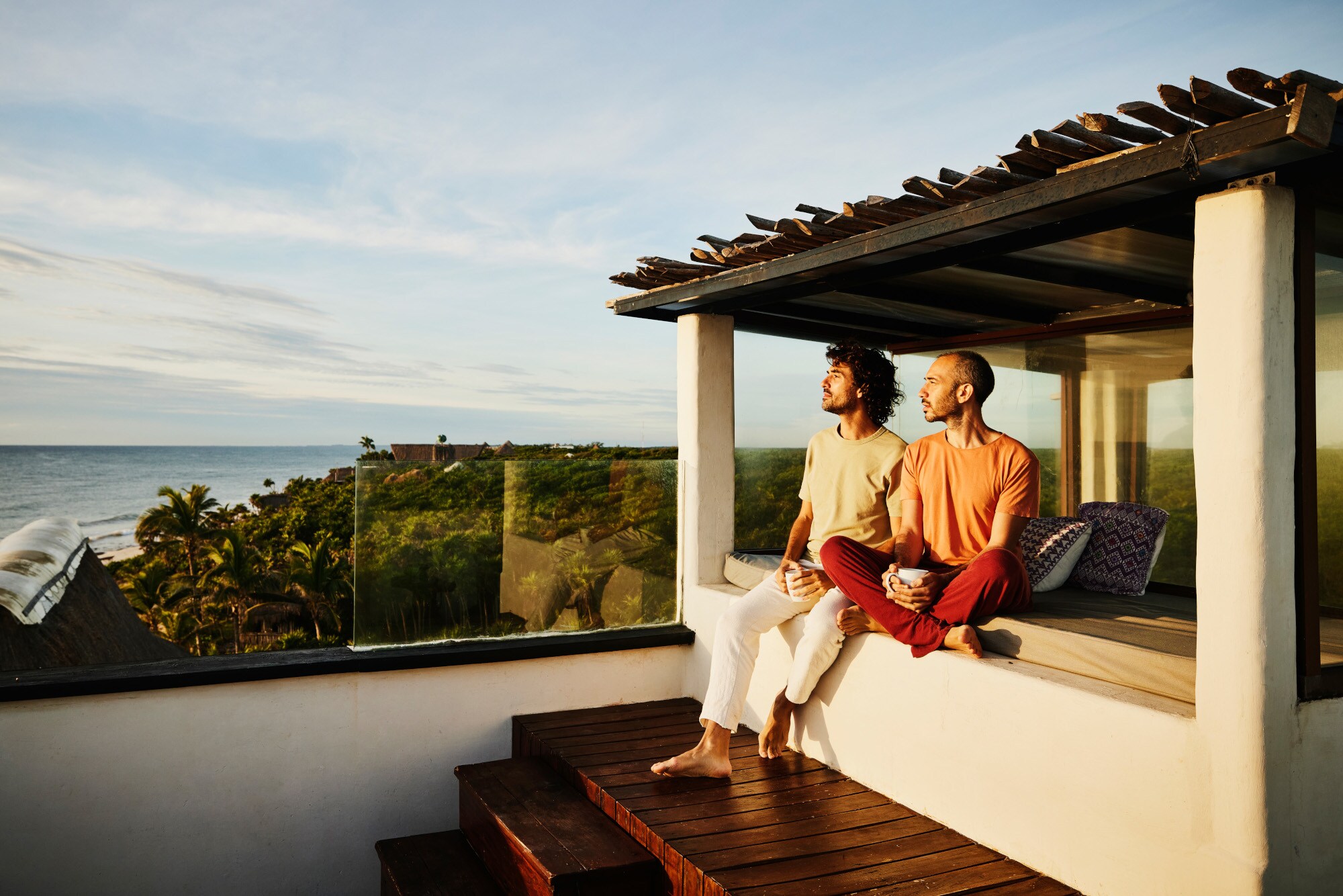 Zwei lässig gekleidete Männer sitzen auf einer Terrasse in der Abendsonne und schauen über eine tropische Landschaft aufs Meer Zwei lässig gekleidete Männer sitzen auf einer Terrasse in der Abendsonne und schauen über eine tropische Landschaft aufs Meer