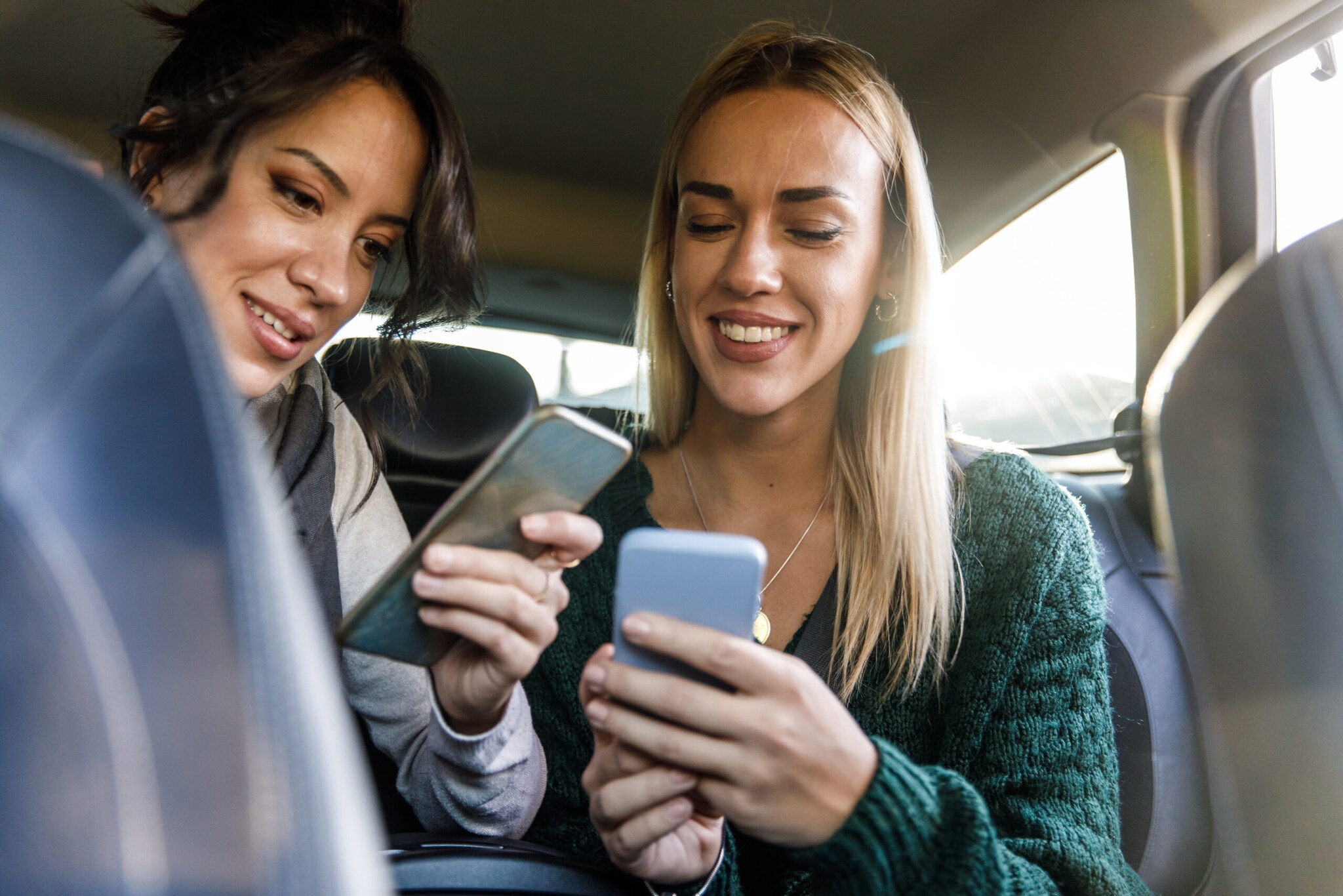 Zwei junge Frauen auf der Rückbank eines Autos schauen lächelnd auf ihre Smartphones