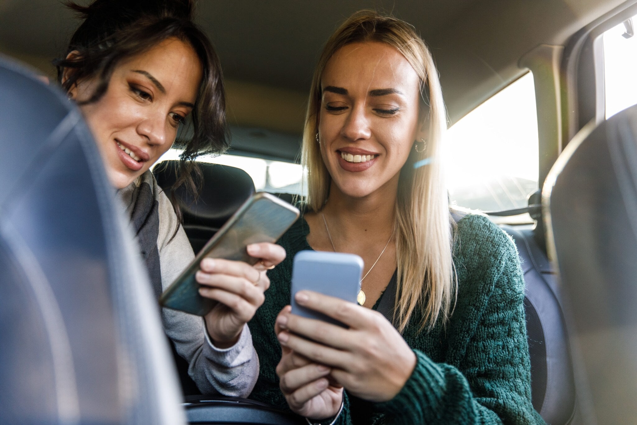 Zwei junge Frauen auf der Rückbank eines Autos schauen lächelnd auf ihre Smartphones Zwei junge Frauen auf der Rückbank eines Autos schauen lächelnd auf ihre Smartphones