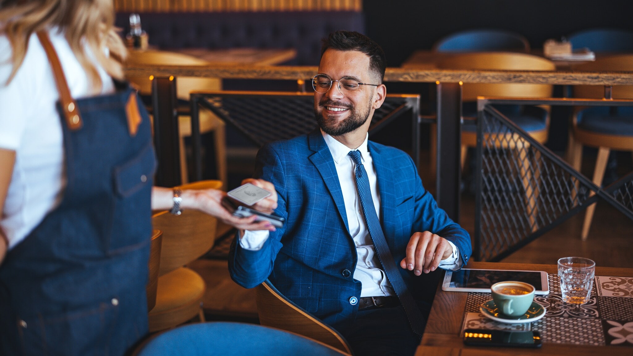 Ein Mann im blauen Anzug sitzt an einem Tisch in einem Bistro und hält eine Kreditkarte an ein Kartenlesegerät, das eine Person ihm hinhält. Ein Mann im blauen Anzug sitzt an einem Tisch in einem Bistro und hält eine Kreditkarte an ein Kartenlesegerät, das eine Person ihm hinhält.