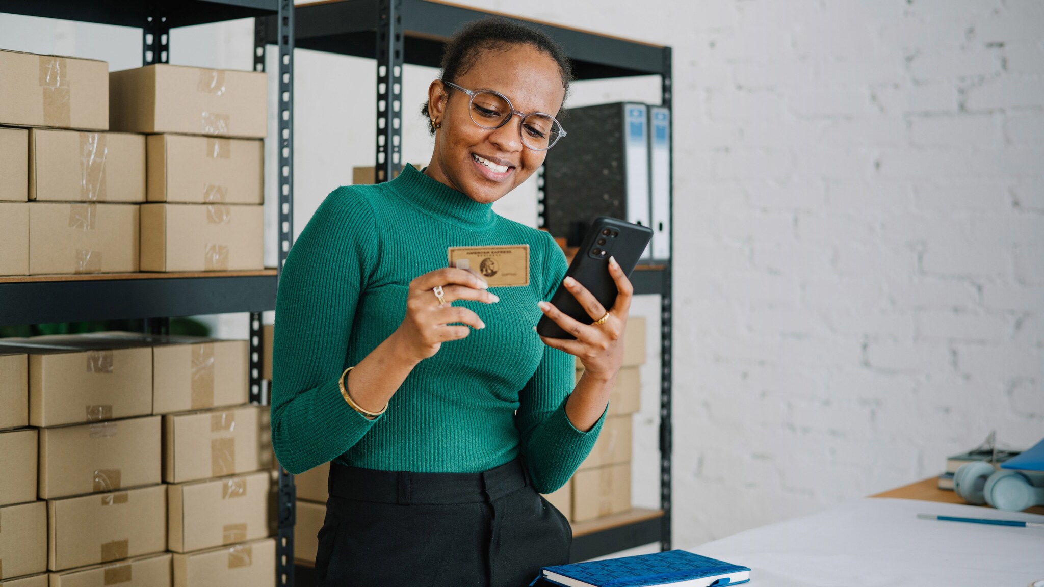 Eine lächelnde Frau im grünen Shirt steht vor einem Lagerregal mit Kartons und hält ein Smartphone sowie eine goldene Kreditkarte von American Express vor sich in den Händen.