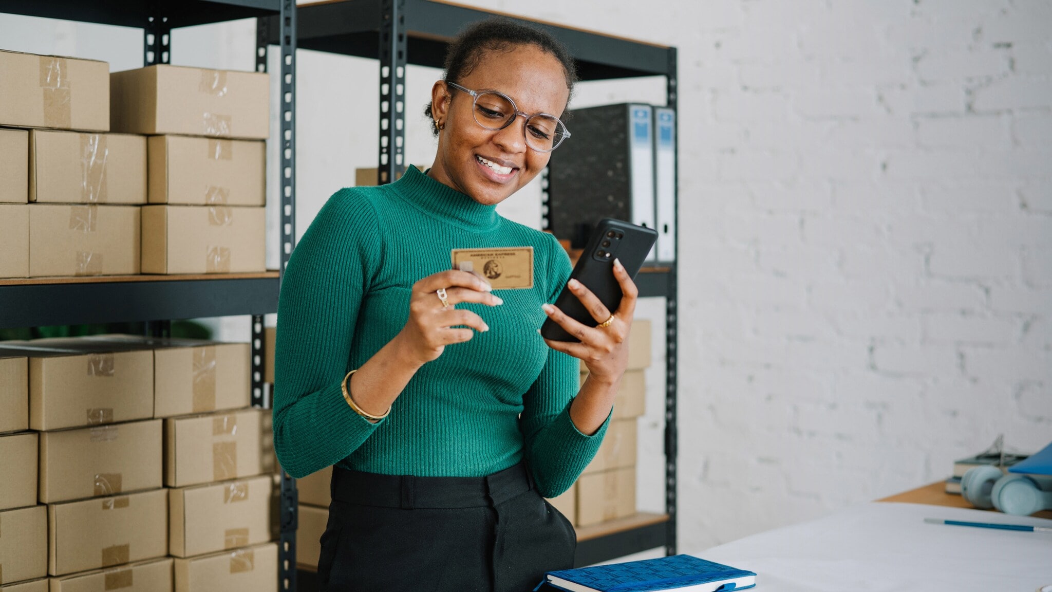 Eine lächelnde Frau im grünen Shirt steht vor einem Lagerregal mit Kartons und hält ein Smartphone sowie eine goldene Kreditkarte von American Express vor sich in den Händen. Eine lächelnde Frau im grünen Shirt steht vor einem Lagerregal mit Kartons und hält ein Smartphone sowie eine goldene Kreditkarte von American Express vor sich in den Händen.
