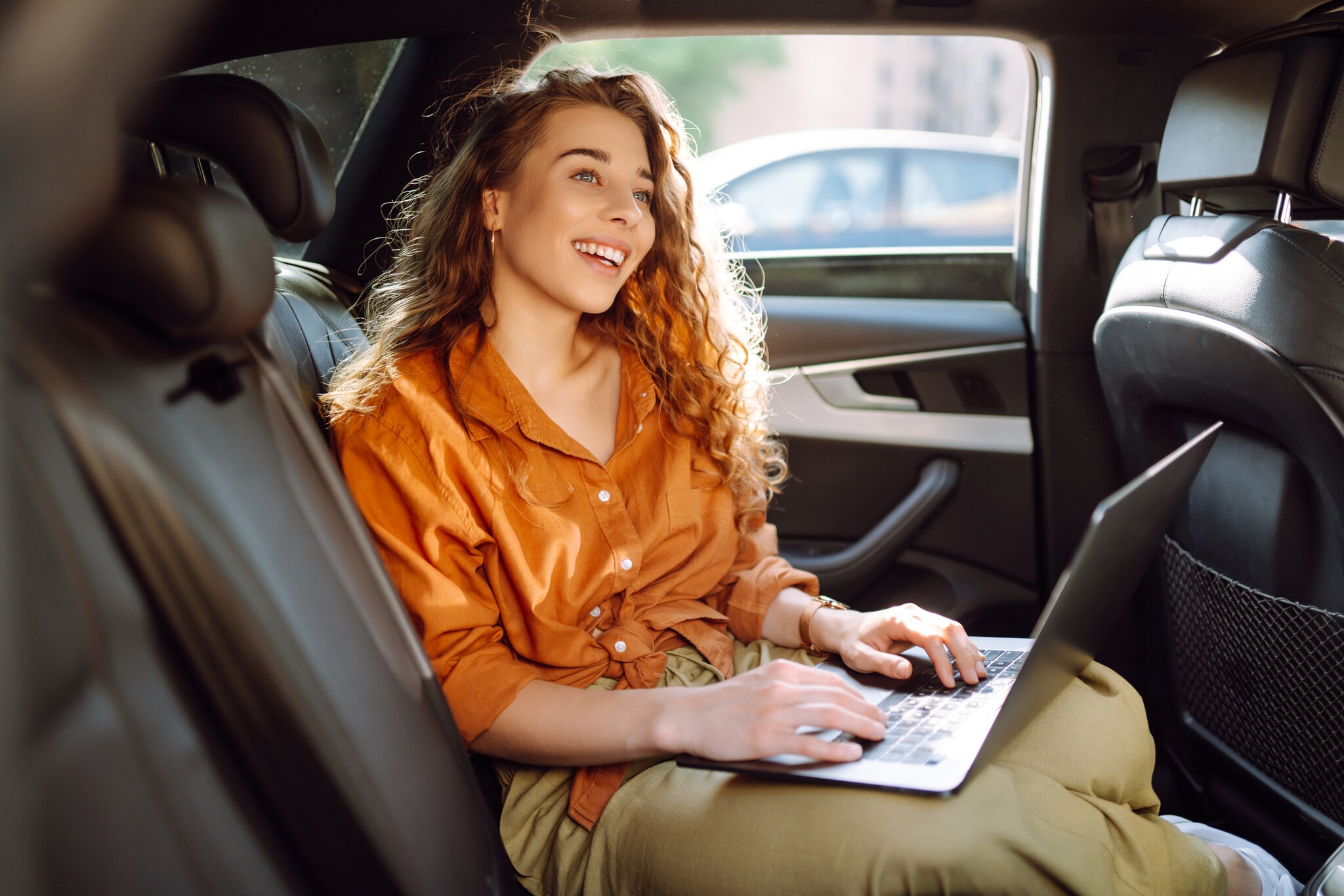 Eine lachende Frau in orangefarbener Bluse sitzt auf dem Rücksitz eines Autos mit einem aufgeklappten Laptop auf ihrem Schoß.