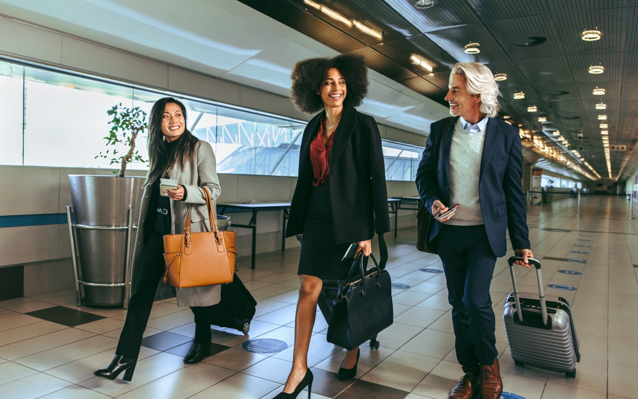 Drei Personen in eleganter Business-Kleidung mit Handgepäck laufen nebeneinander durch ein Flughafenterminal. Drei Personen in eleganter Business-Kleidung mit Handgepäck laufen nebeneinander durch ein Flughafenterminal.