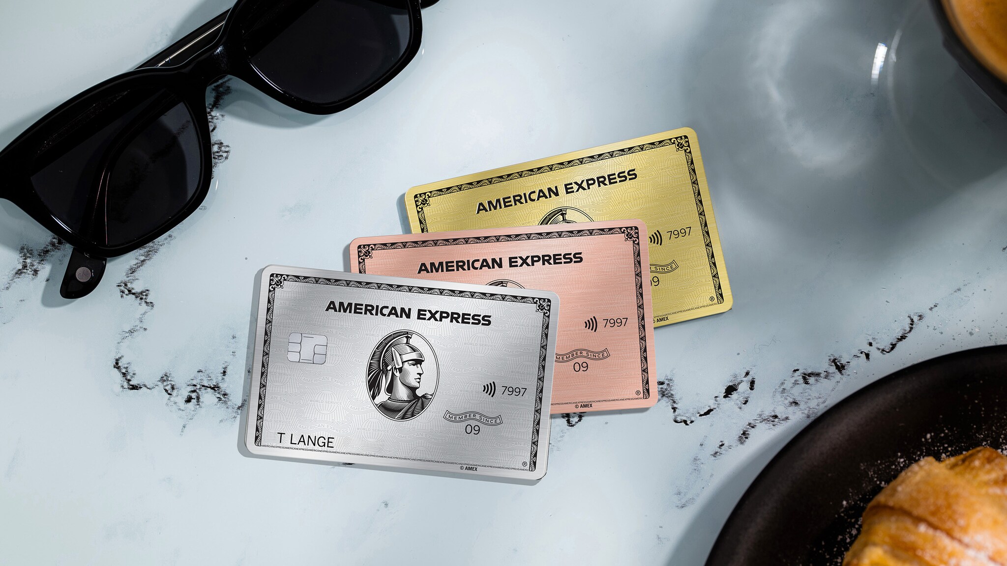 Drei Kreditkarten von American Express liegen auf einem Kaffeetisch aus Marmor, drum herum ist ein Croissants und eine Sonnenbrille.