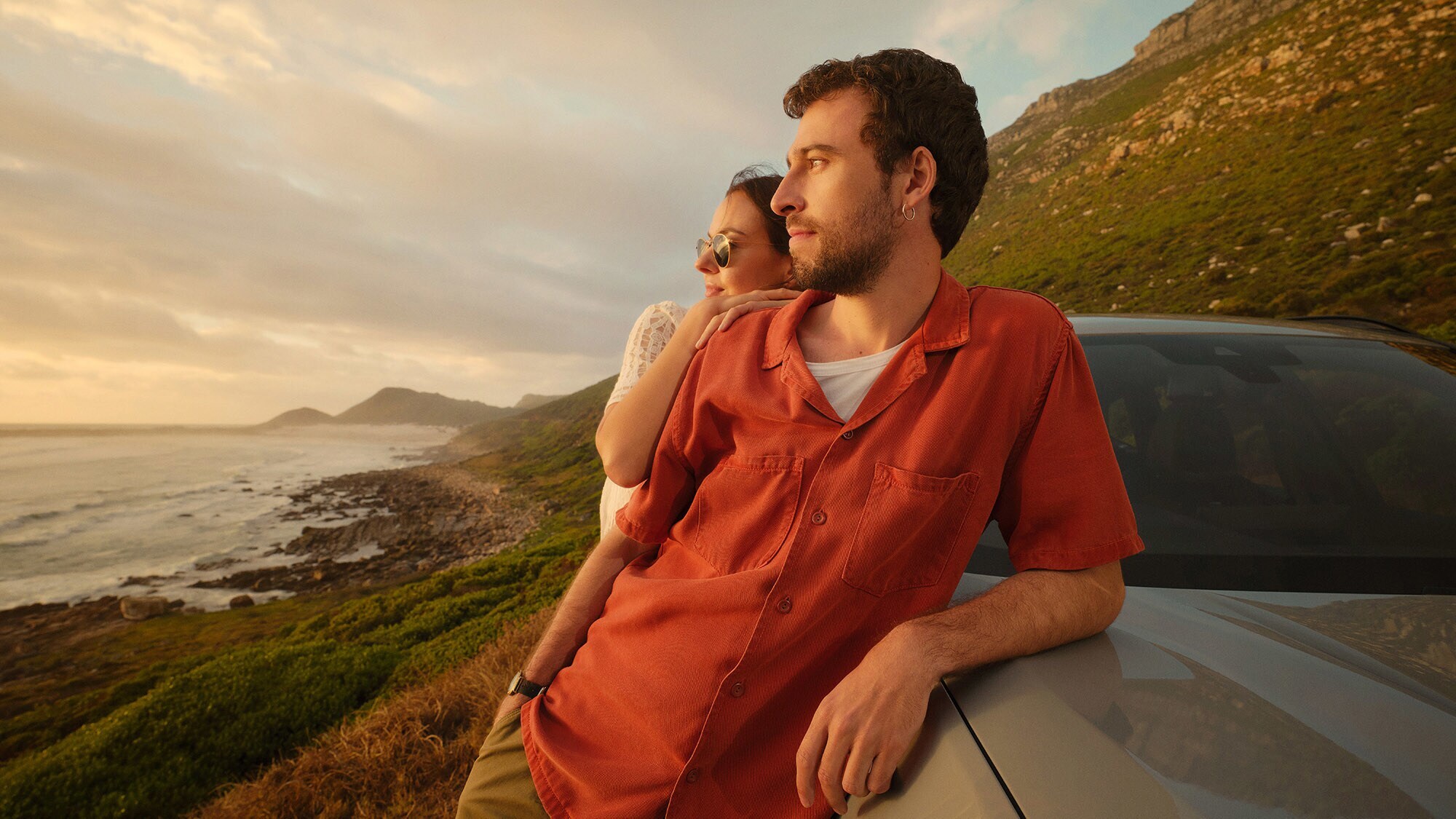 Ein Paar lehnt seitlich an einem Auto an einer Küste und blickt aufs Meer.