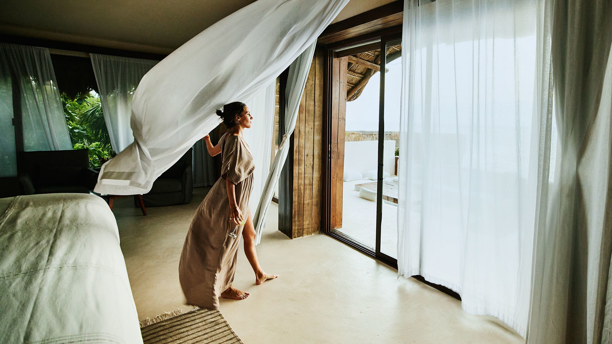 Eine Frau im langen Kleid und einem Weinglas in der Hand steht in einer Suite und blickt hinaus durch eine geöffnete Terassentür mit wehenden Gardinen. Eine Frau im langen Kleid und einem Weinglas in der Hand steht in einer Suite und blickt hinaus durch eine geöffnete Terassentür mit wehenden Gardinen.