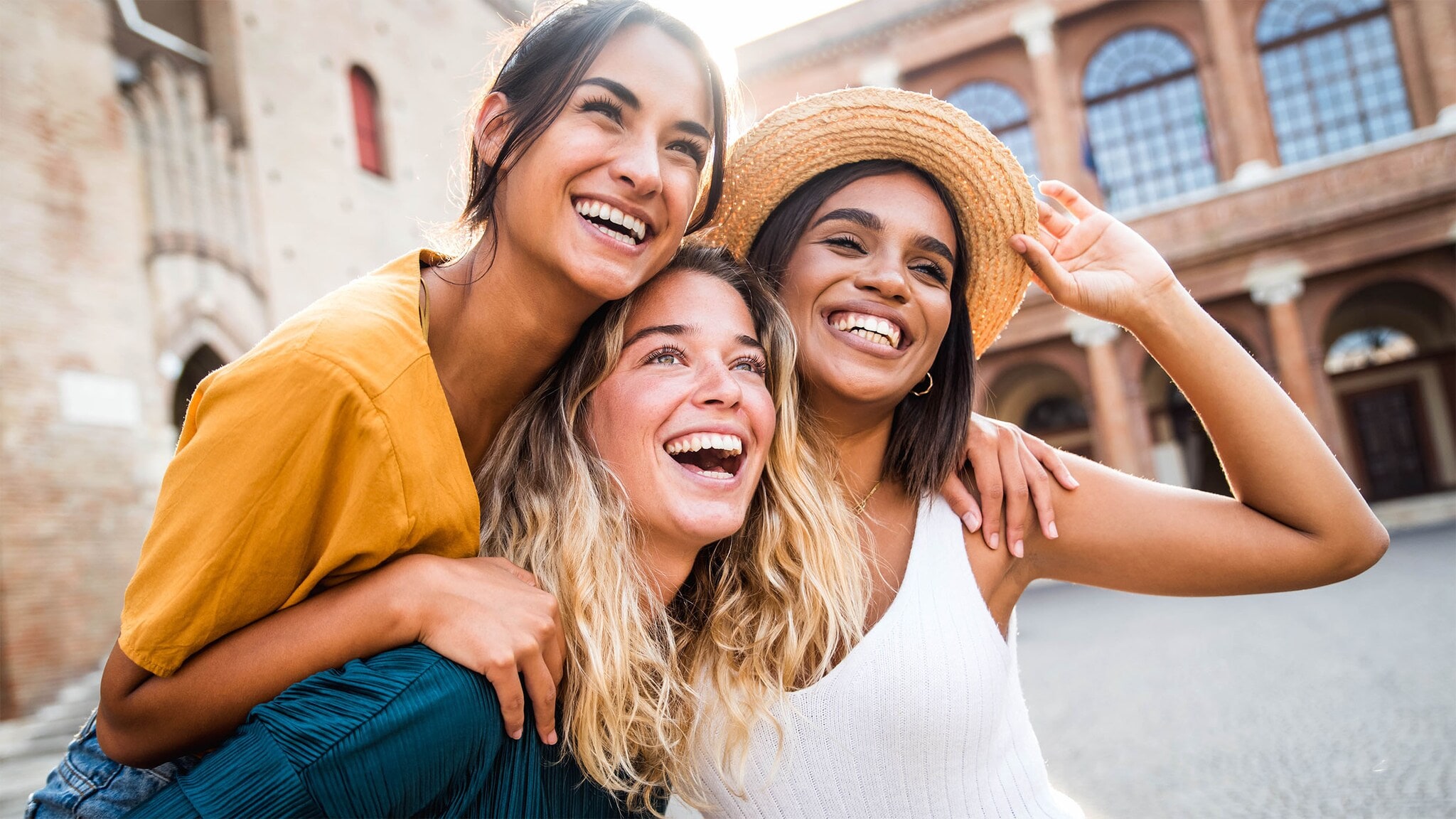 American Express MGM Drei lachende junge Frauen stehen sich umarmend auf einem Platz.