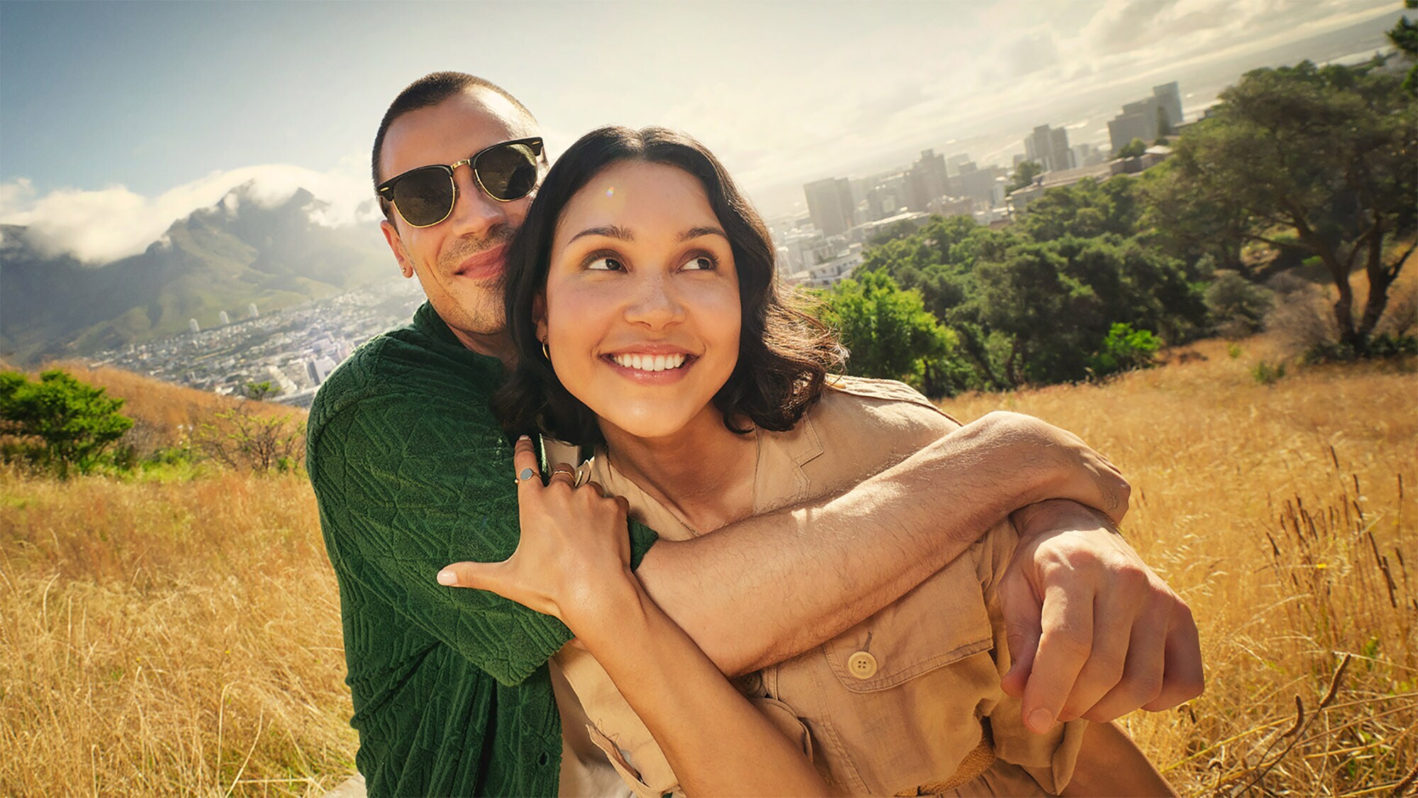 Ein sich umarmendes, junges Paar auf einem Hügel vor der Skyline von Kapstadt.
