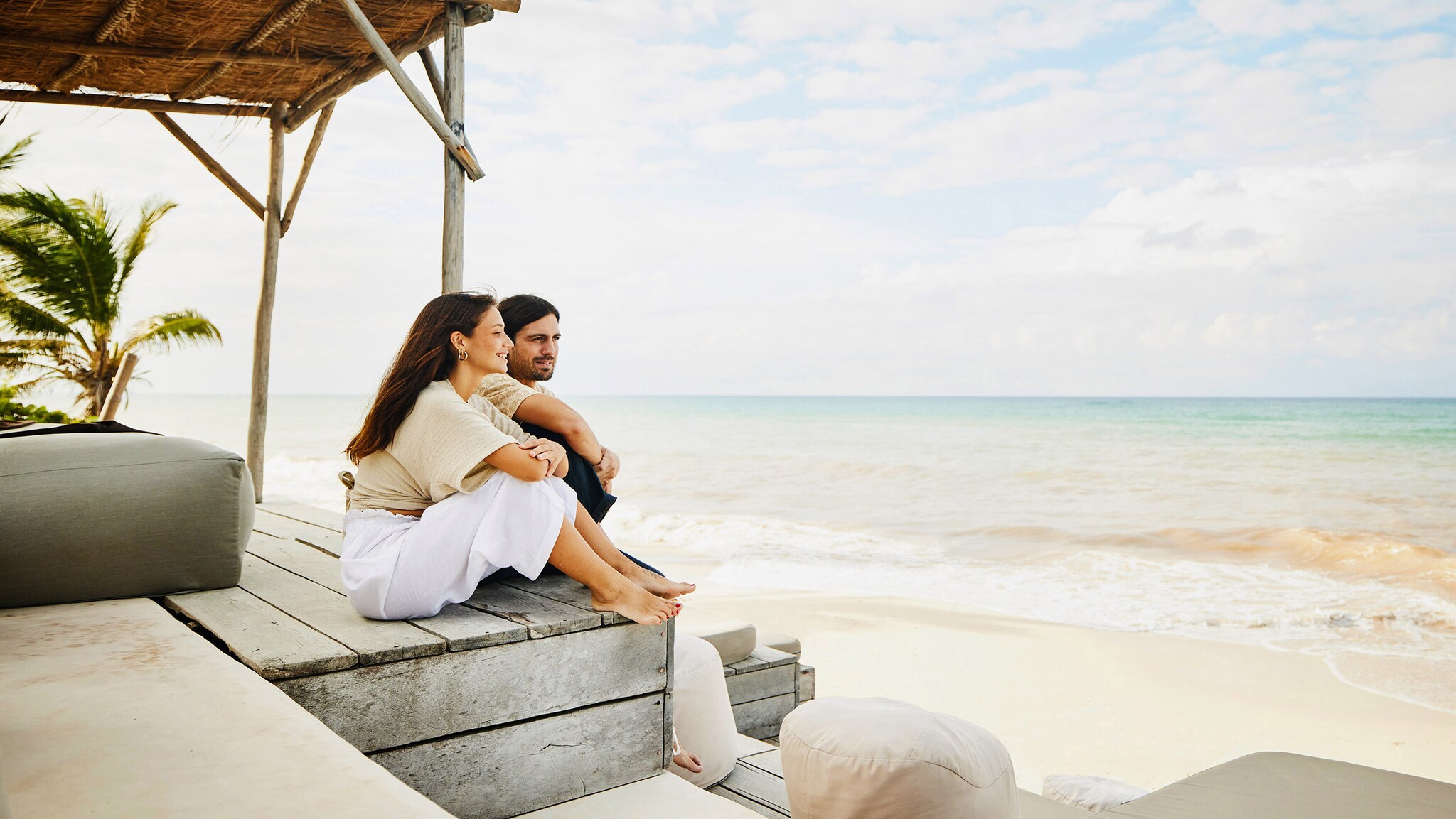 Ein Paar sitzt entspannt auf einer Holztribüne mit Polstermöbeln an einem weißen Sandstrand und blickt aufs Meer.