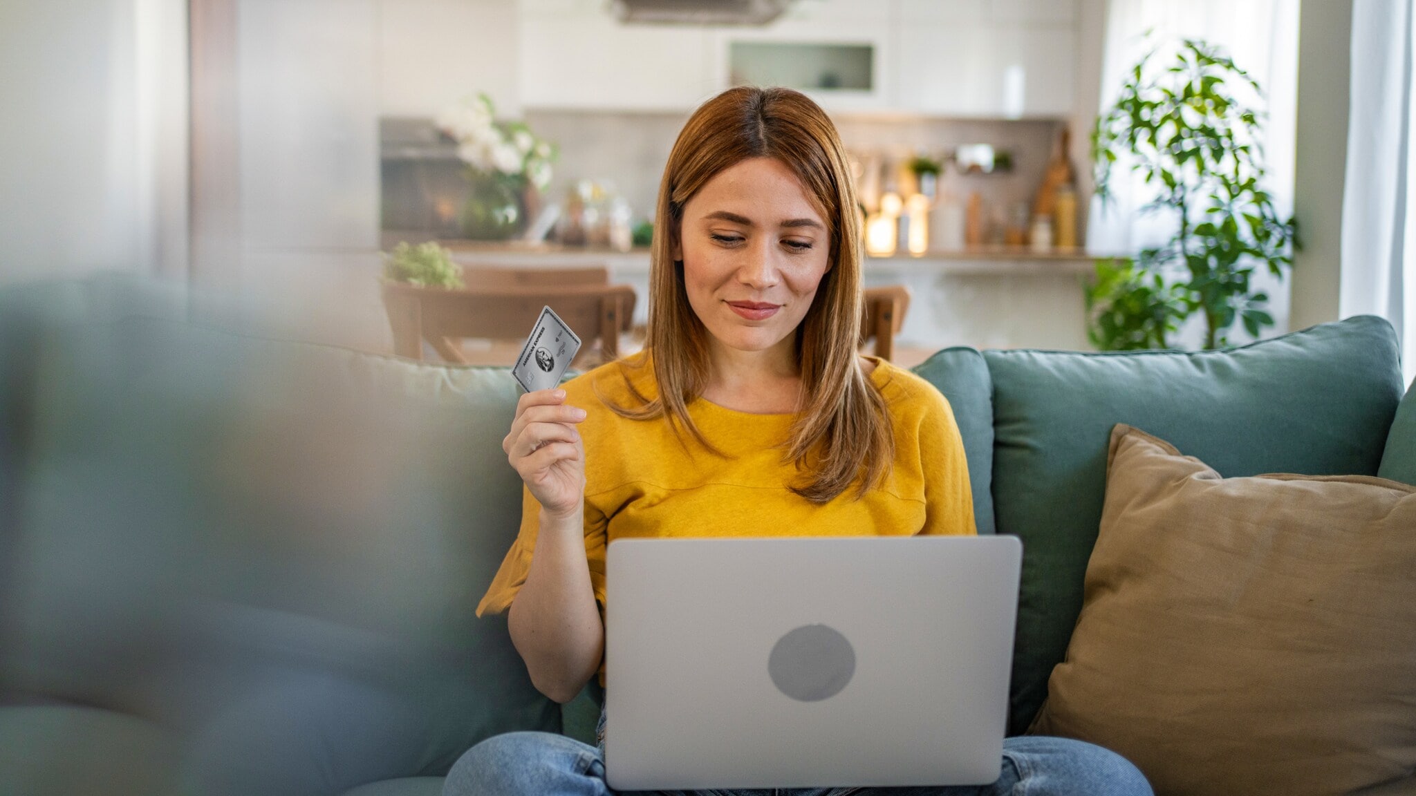 Eine junge Frau im gelben T-Shirt sitzt mit einem Laptop auf dem Schoß auf einem Sofa und schaut lächelnd auf das Display, in ihrer Hand hält sie eine silberne Kreditkarte von American Express. Eine junge Frau im gelben T-Shirt sitzt mit einem Laptop auf dem Schoß auf einem Sofa und schaut lächelnd auf das Display, in ihrer Hand hält sie eine silberne Kreditkarte von American Express.