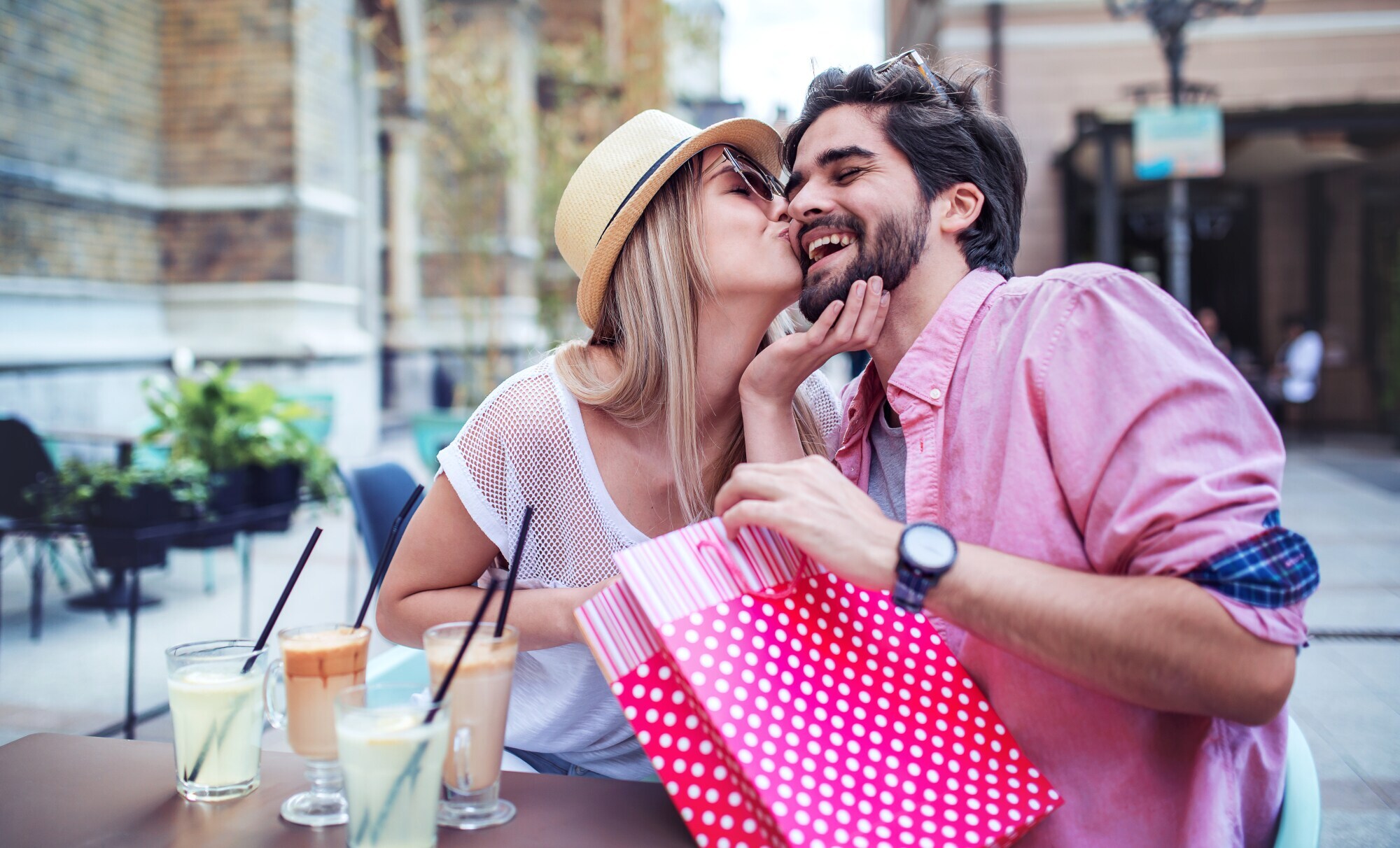 Ein junges, glückliches Paar mit einer roten Geschenktüte an einem Tisch im Außenbereich eines Cafés, sie gibt ihm einen Kuss auf die Wange. Ein junges, glückliches Paar mit einer roten Geschenktüte an einem Tisch im Außenbereich eines Cafés, sie gibt ihm einen Kuss auf die Wange.