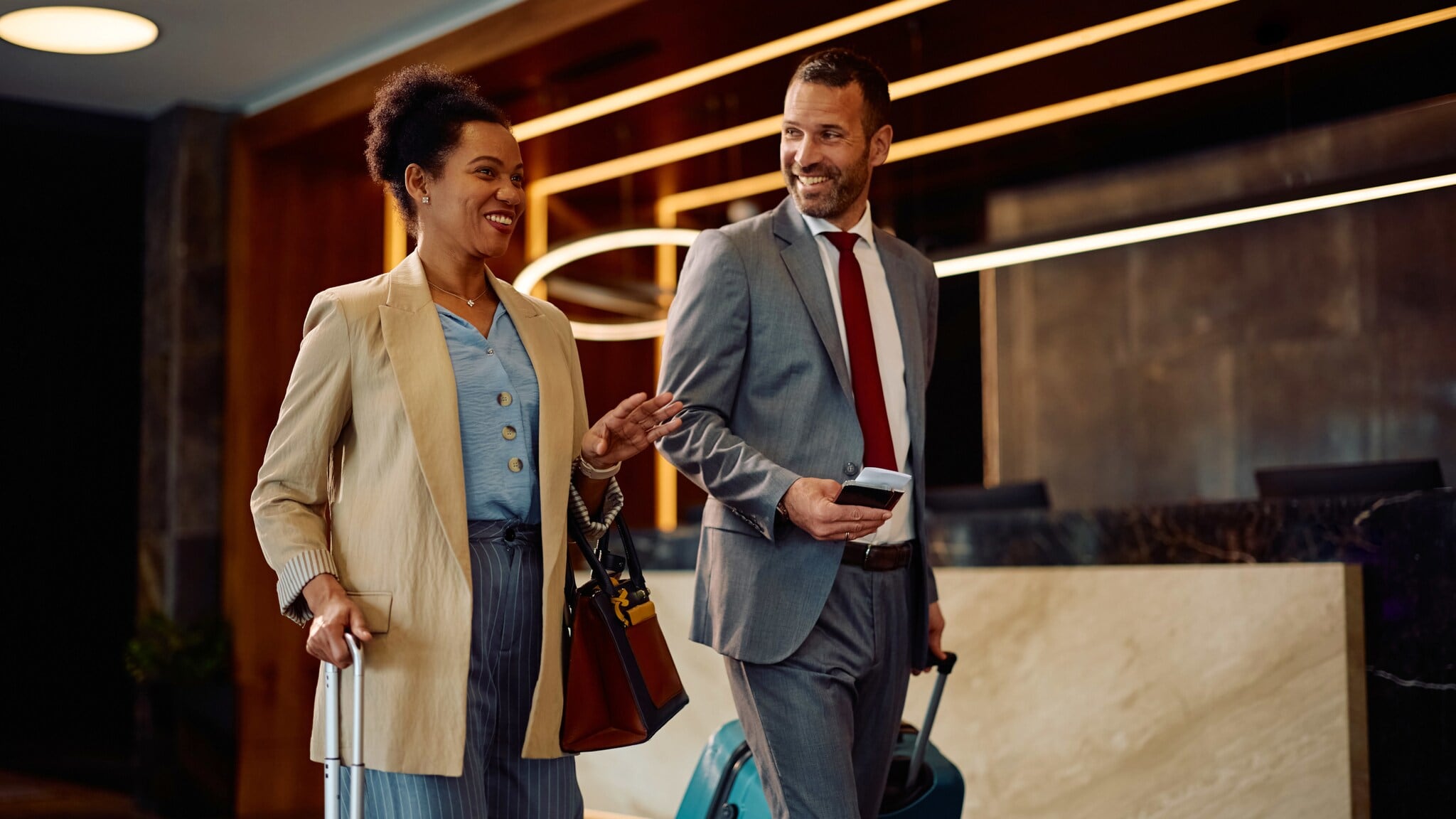Eine Frau und ein Mann in Business-Kleidung mit Trolleys gehen gut gelaunt durch eine Hotellobby. Eine Frau und ein Mann in Business-Kleidung mit Trolleys gehen gut gelaunt durch eine Hotellobby.