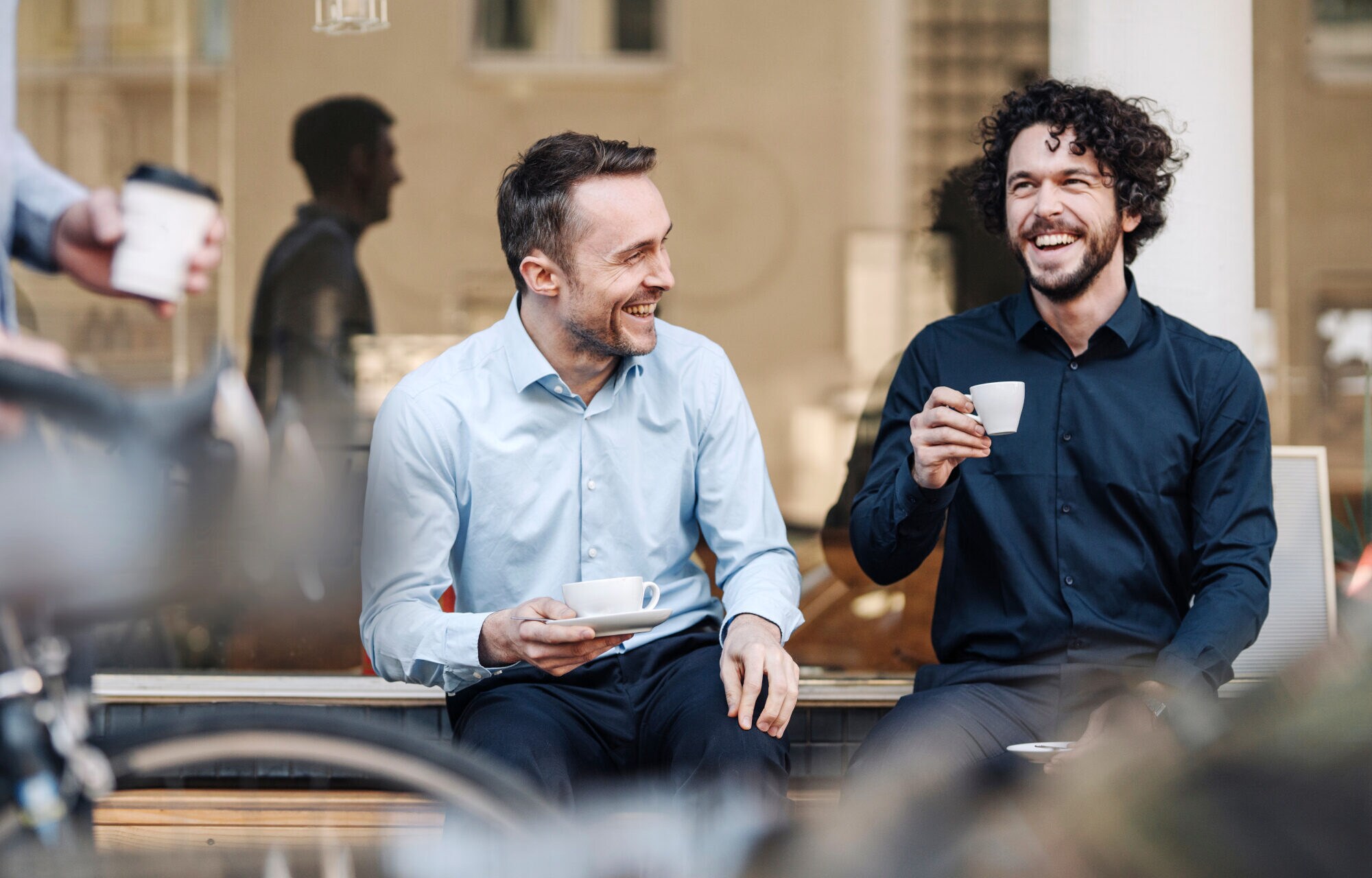 Zwei Männer genießen gut gelaunt einen Kaffee auf einer Bank vor einem Café