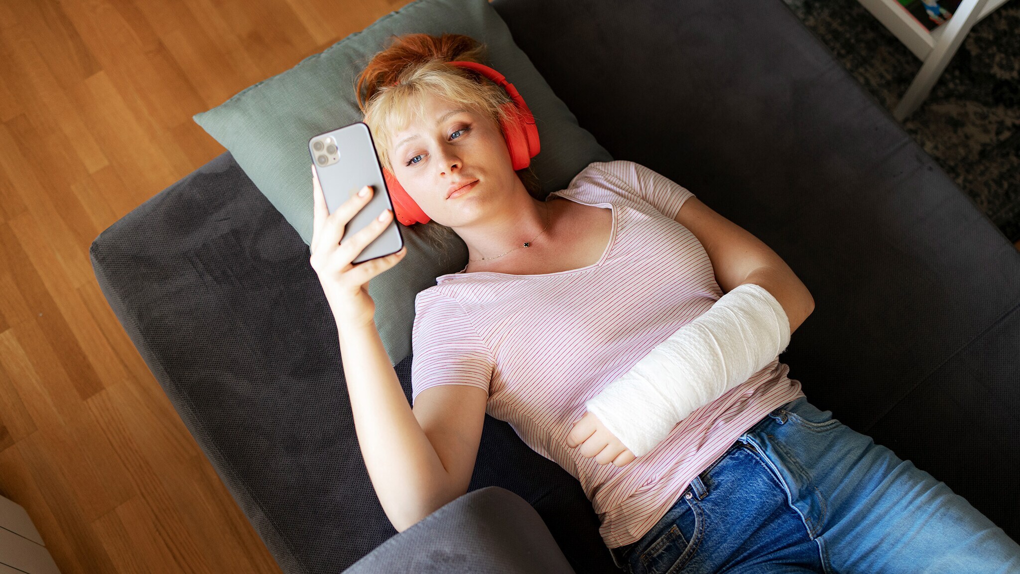 Aufsicht einer auf einem Sofa liegenden, jungen Frau, die mit Kopfhörern und einem eingegipsten Arm auf ihr Smartphone schaut.