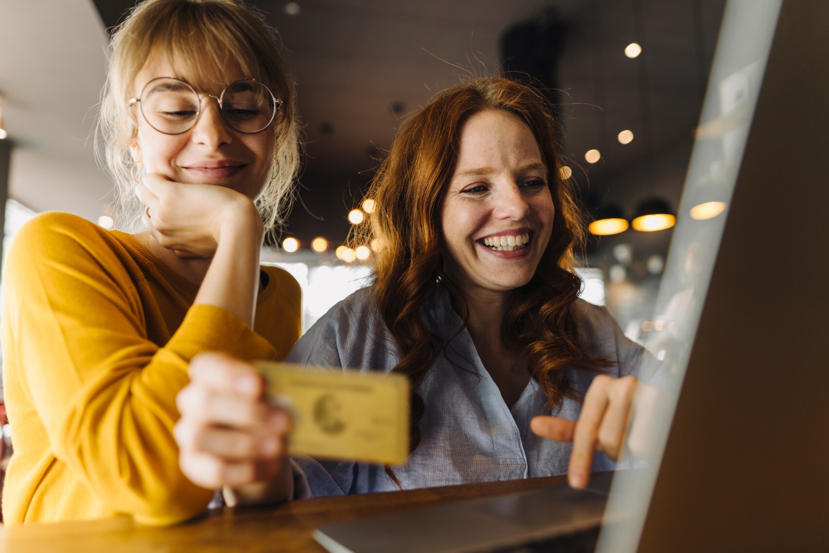 Zwei junge Frauen sitzen lächelnd mit einer Kreditkarte vor einem Laptop in einem Café.
