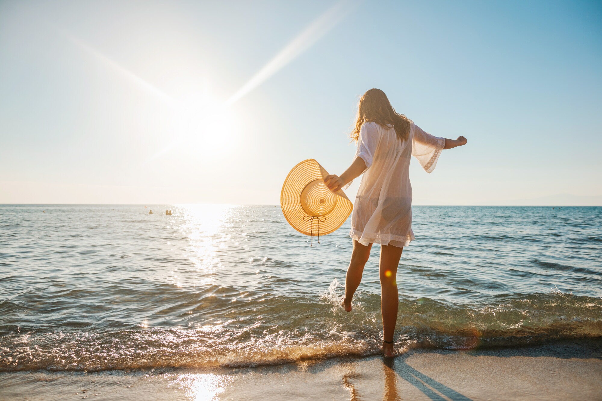 Eine junge Frau im weißen Sommerkleid und mit Strohhut in der Hand planscht mit ihrem Fuß im Wasser an einem Sandstrand am Meer bei Sonnenuntergang