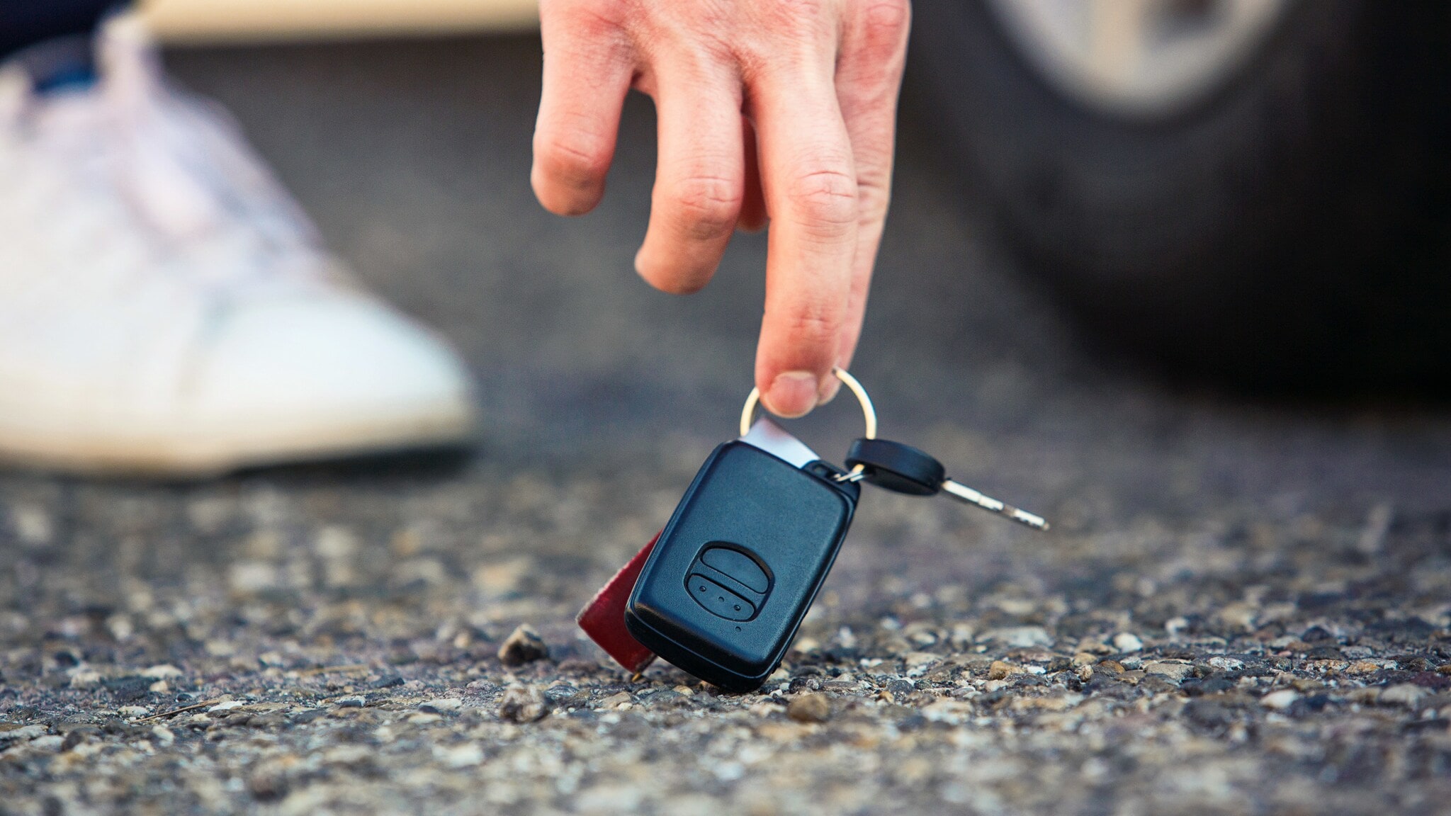 Nahaufnahme einer Hand, die nach einem Autoschlüssel am Asphaltboden greift, dahinter ein Autoreifen und Sneaker in Unschärfe. Nahaufnahme einer Hand, die nach einem Autoschlüssel am Asphaltboden greift, dahinter ein Autoreifen und Sneaker in Unschärfe.
