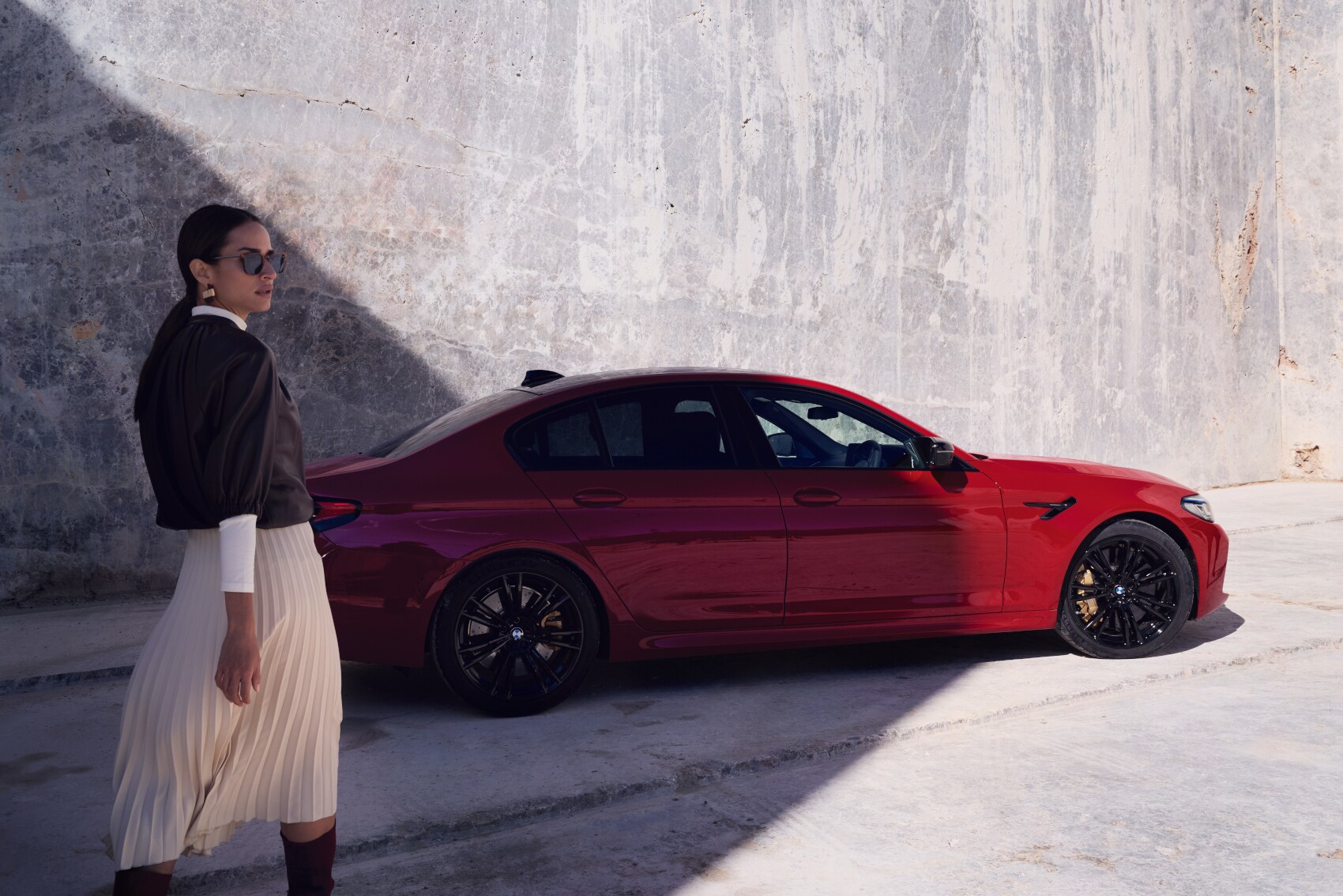 Eine elegante Frau mit Sonnenbrille vor einem roten BMW vor einer Betonwand.