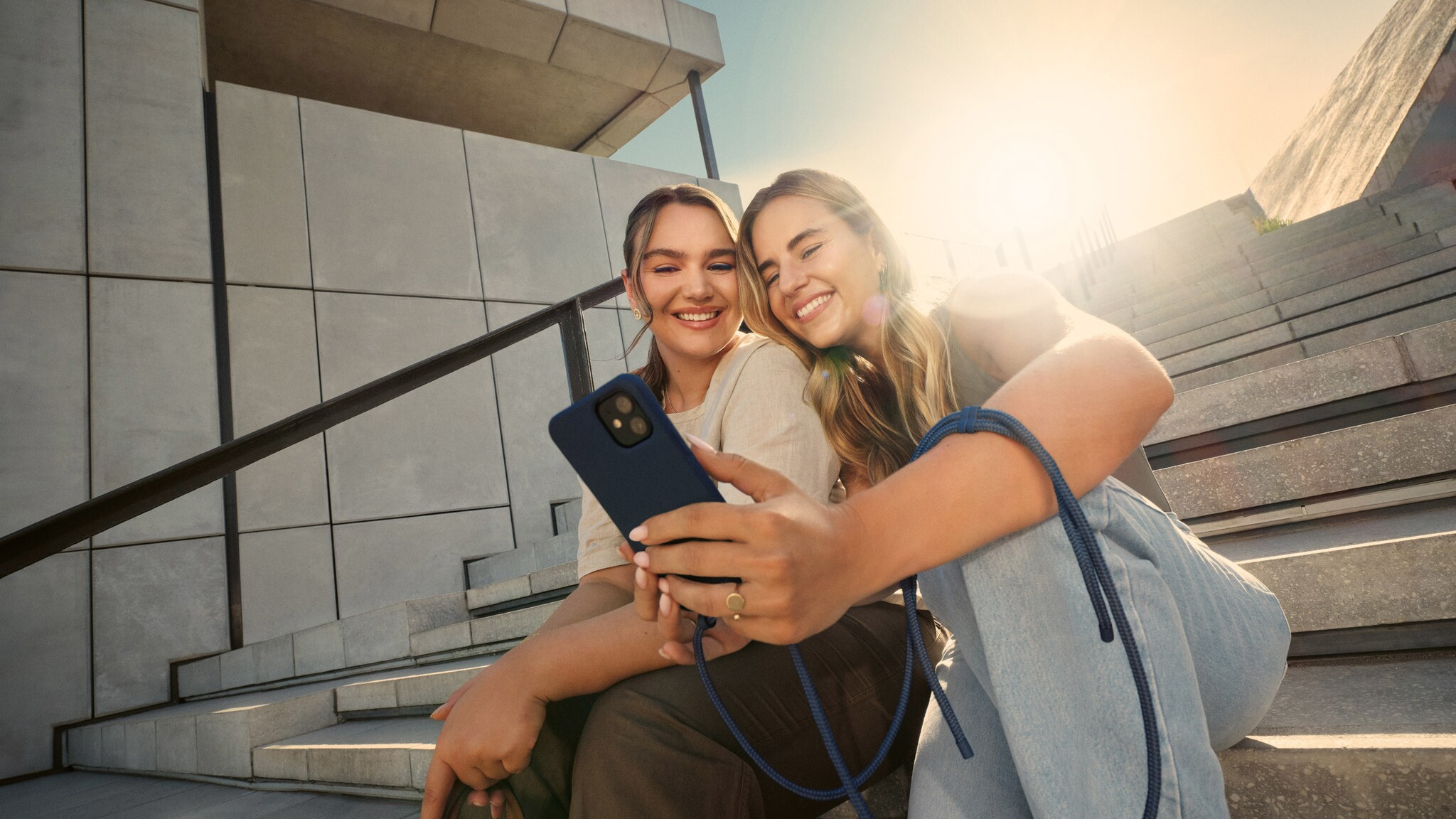Zwei lächelnde junge Frauen sitzen auf einer Treppe im Freien und schauen gemeinsam auf ein Smartphone. Zwei lächelnde junge Frauen sitzen auf einer Treppe im Freien und schauen gemeinsam auf ein Smartphone.