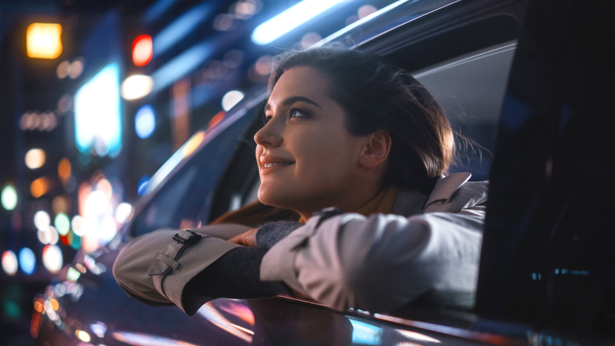 Nahaufnahme einer lächelnden Frau, die von der Rückbank eines fahrenden Autos aus dem geöffneten Fenster blickt, im Hintergrund Leuchtreklame in urbaner Umgebung bei Nacht. Nahaufnahme einer lächelnden Frau, die von der Rückbank eines fahrenden Autos aus dem geöffneten Fenster blickt, im Hintergrund Leuchtreklame in urbaner Umgebung bei Nacht.