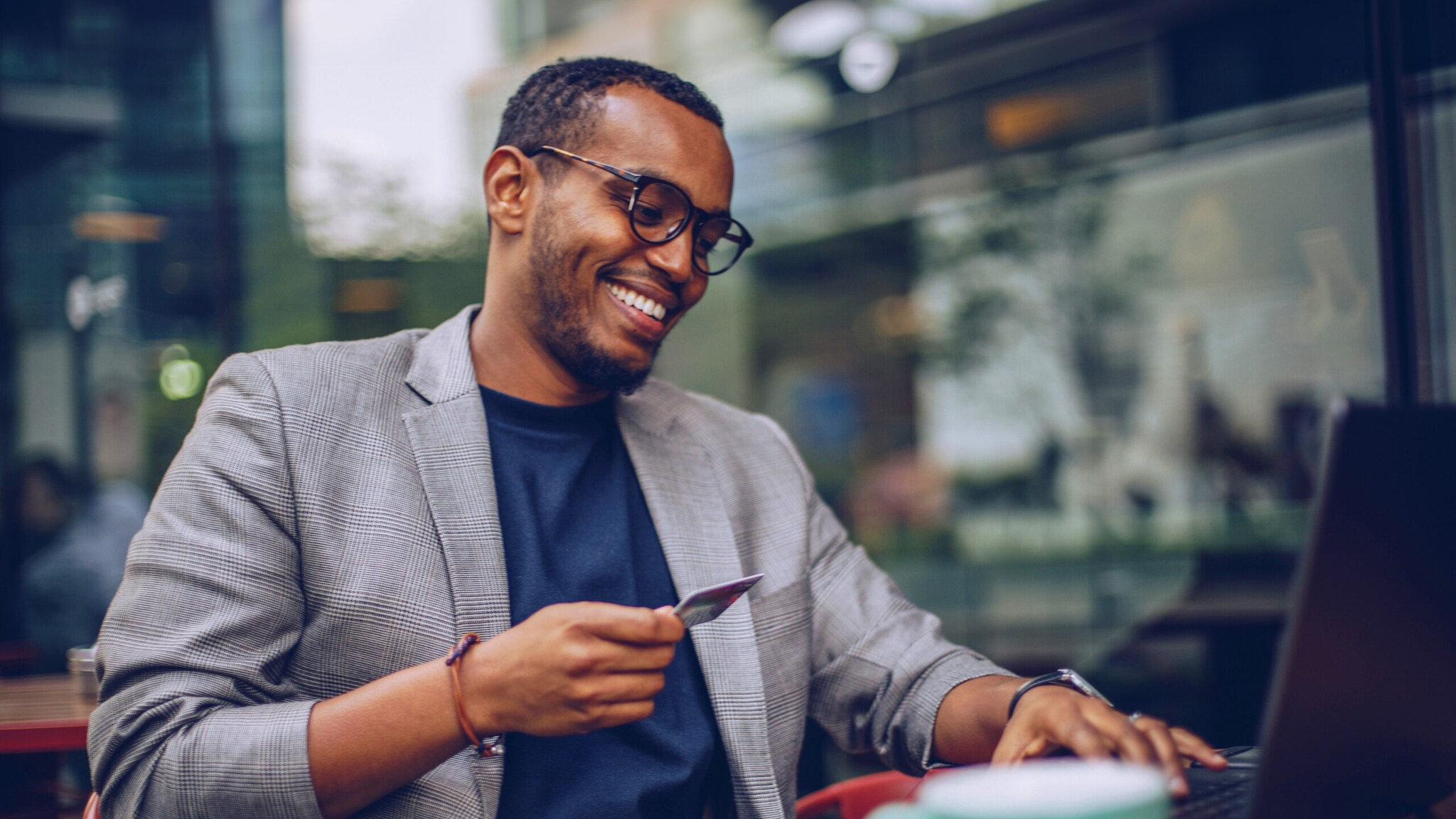 Ein junger, lächelnder Mann sitzt mit einer Kreditkarte in der Hand vor einem Laptop in einem Café.