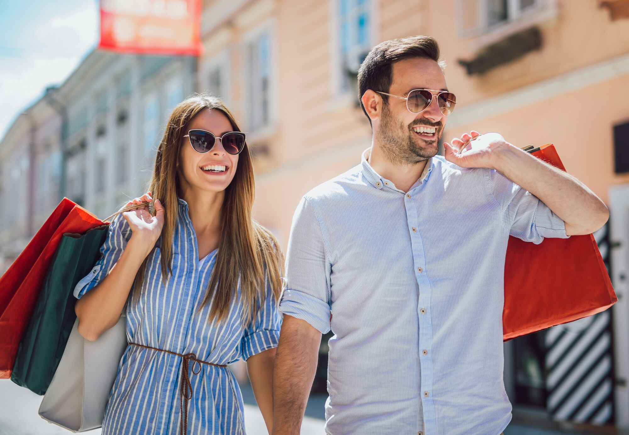 Ein lachendes Paar mit Sonnenbrillen und Einkaufstüten auf einer Straße.