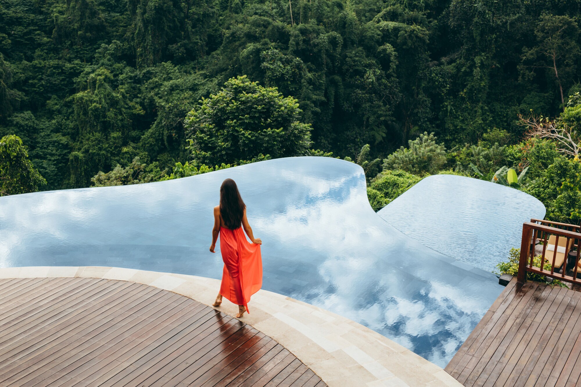 Rückansicht einer Frau im langen Sommerkleid an einem Infinitypool auf einer Terrasse, umgeben von tropischem Regenwald.